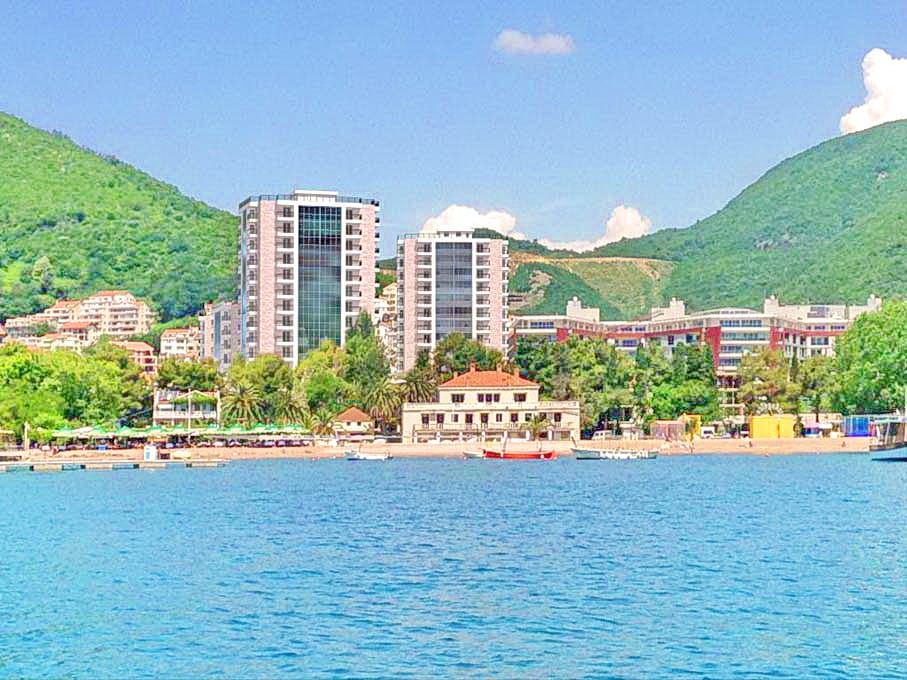 Pogled sa mora na moderne stambene komplekse i plažu u gradu Budvi, Crna Gora, u pozadini zelenih planina i plavog neba.