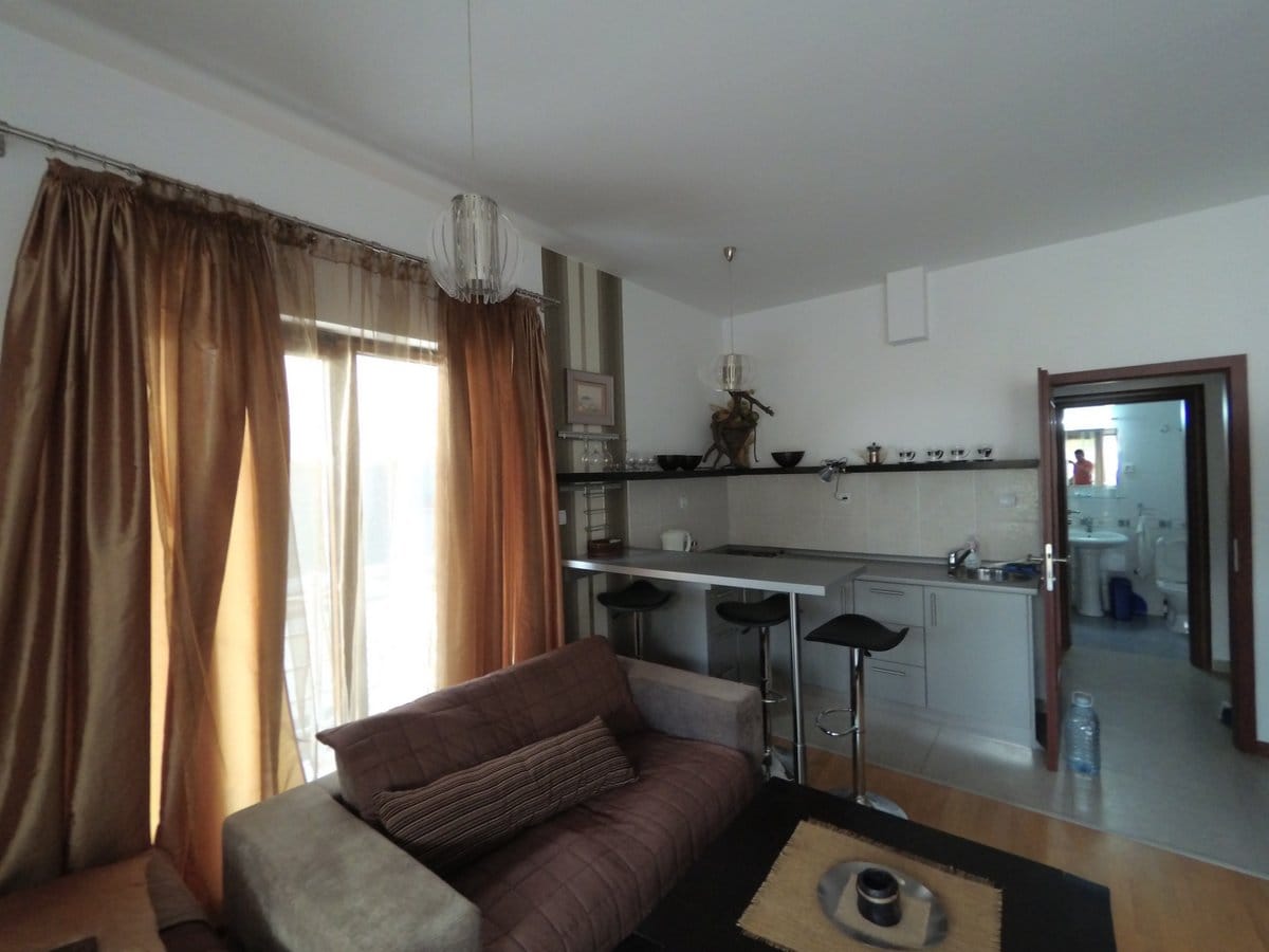 Dnevna soba sa kuhinjom i barskim pultom u stanu u Budvi, Crna Gora: savremeni nameštaj, svetla završna obrada i izlaz na balkon.
