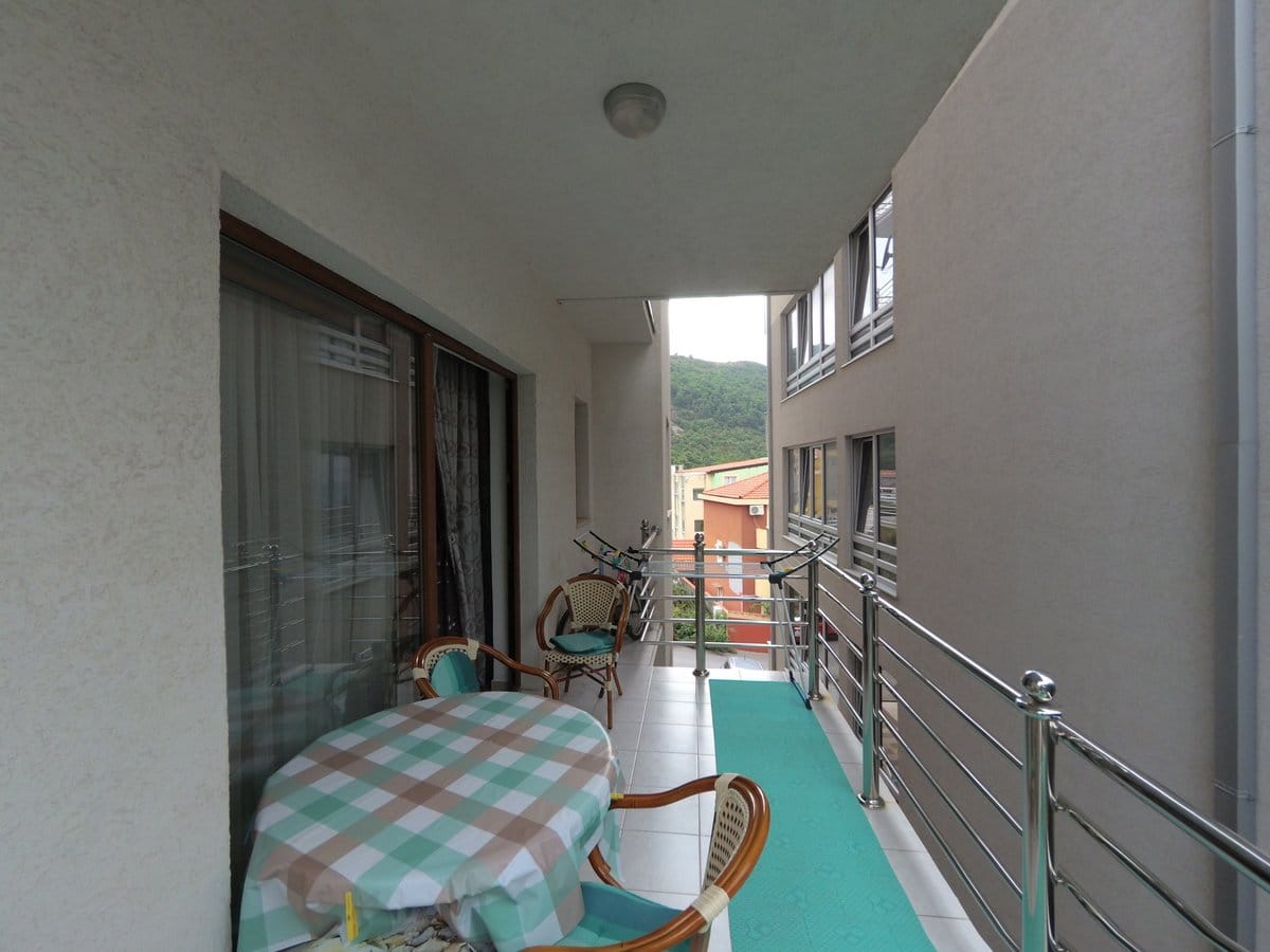 Spacious balcony with wicker furniture in an apartment in Budva, Montenegro, overlooking neighboring houses and green mountains.