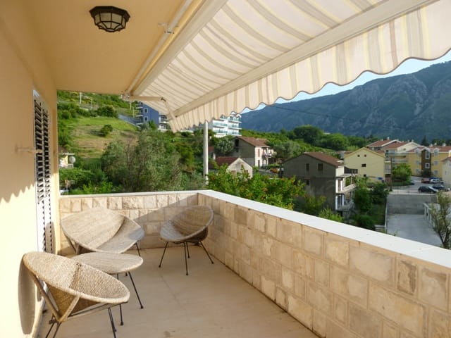 Ugodan balkon s pletenim nameštajem i nadstrešnicom na vili u Dobroti, Crna Gora, s panoramskim pogledom na planine i grad.