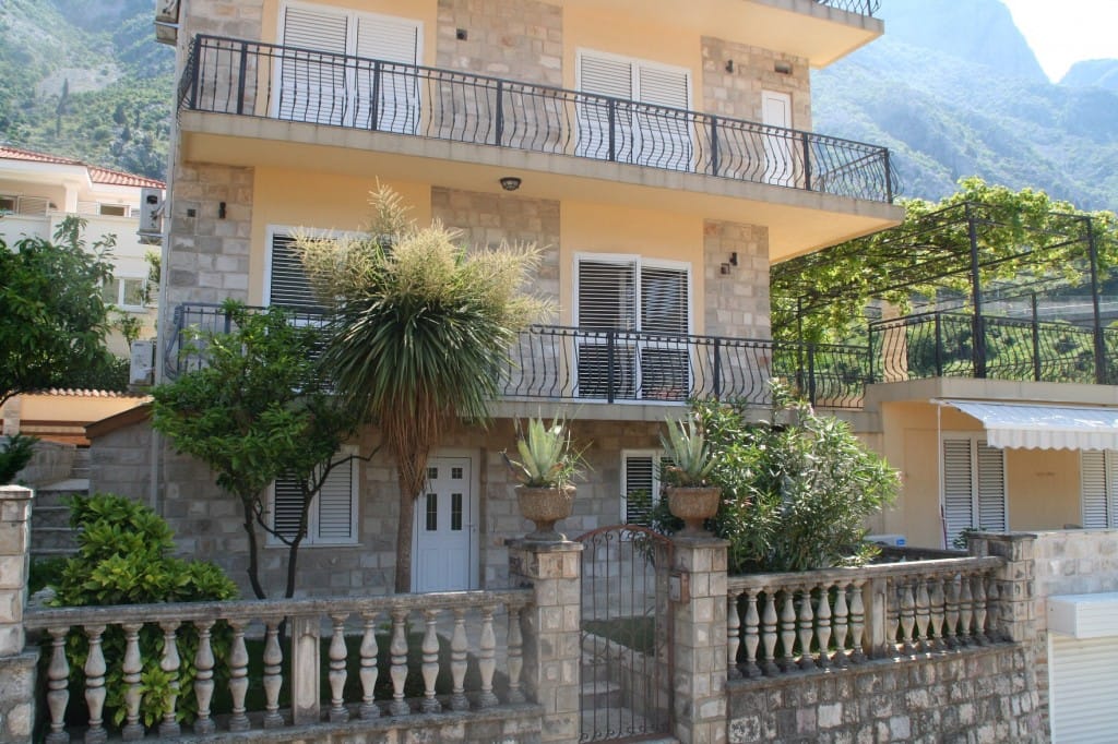 Trospratna vila u Dobroti, Crna Gora: kamena fasadna obloga, kovani balkoni, bašta sa palmom i pogled na planine.