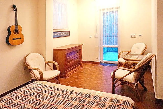 Cozy bedroom in an apartment in Stoliv, Montenegro: wicker furniture, a wooden dresser, a guitar on the wall, and access to the balcony.