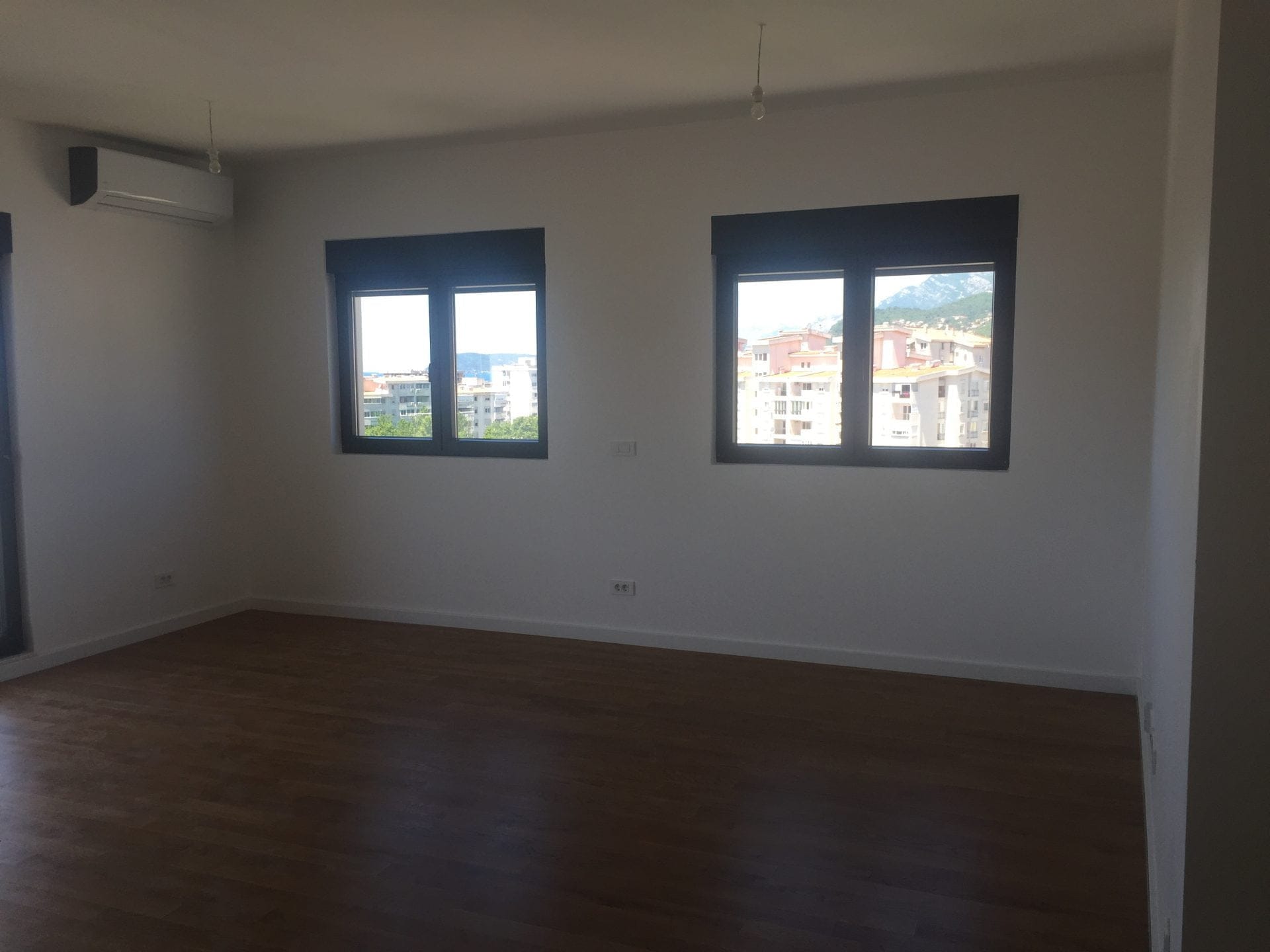 Helles Wohnzimmer mit Parkettboden und Klimaanlage in einer Wohnung in der Stadt Bar, Montenegro, mit Blick auf die Stadt und die Berge aus den Fenstern.