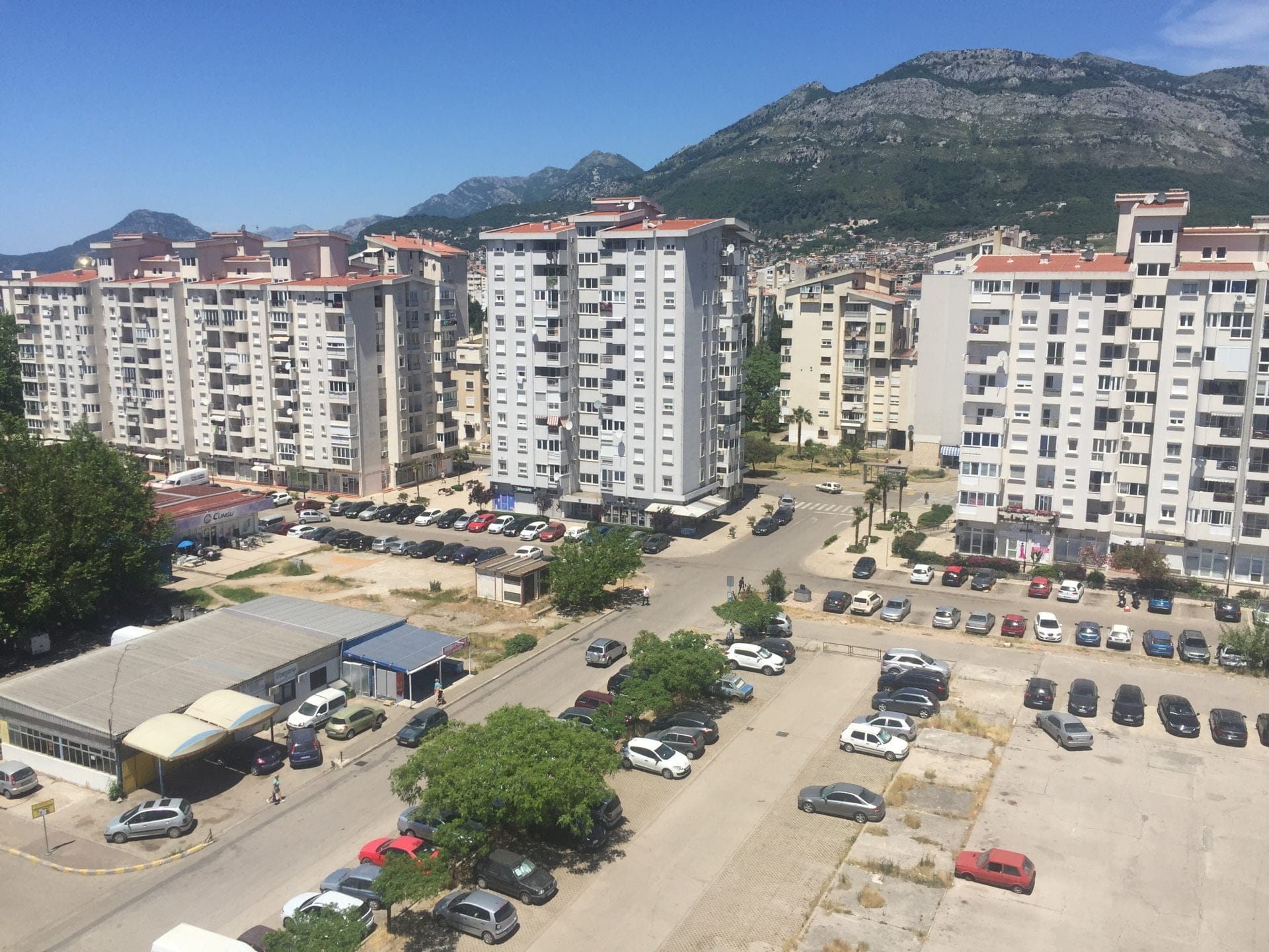 Blick aus dem Fenster auf ein Wohnviertel und Berge in der Stadt Bar, Montenegro: mehrstöckige Wohnhäuser und ein Parkplatz mit Autos.