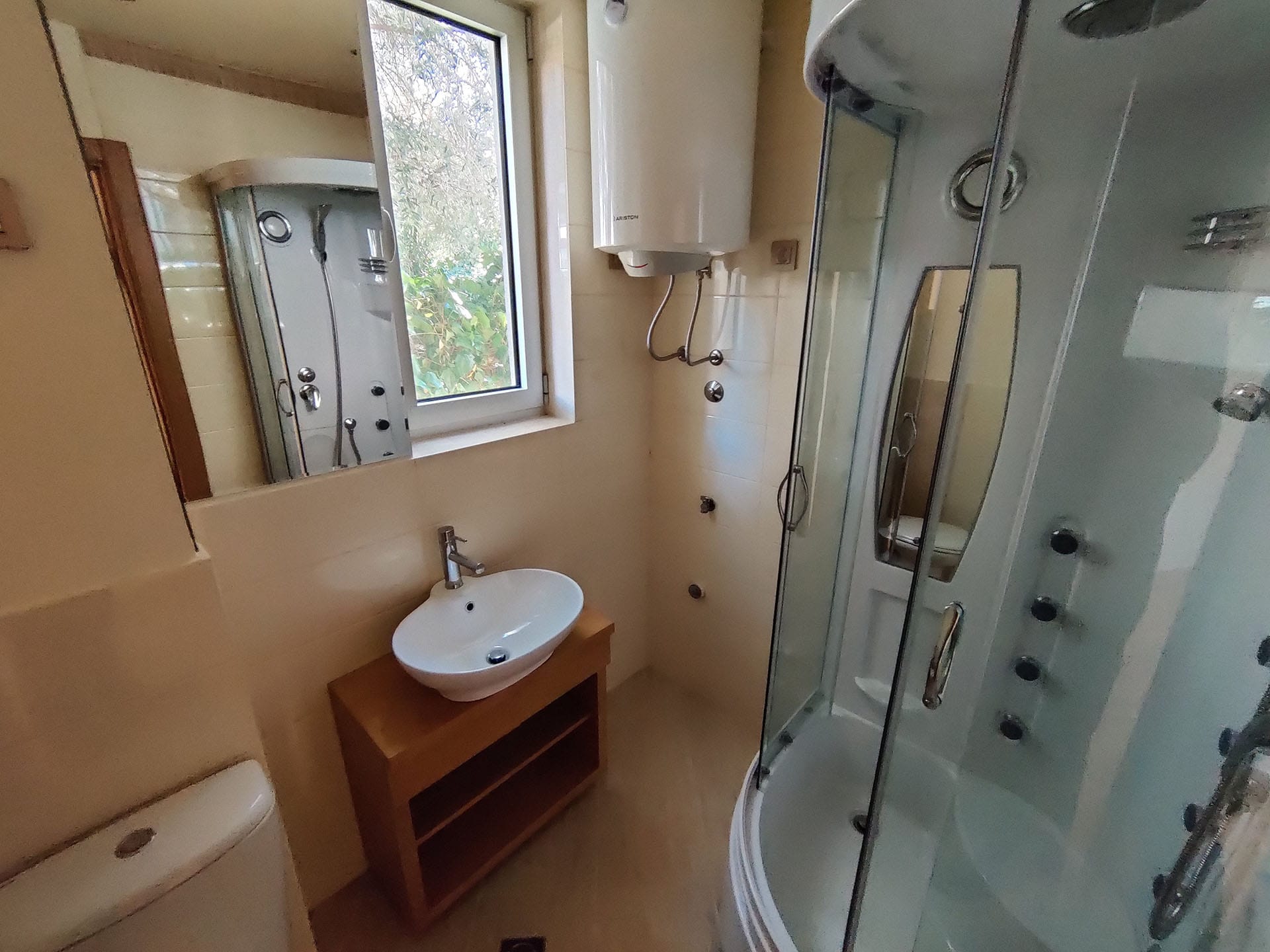 Salle de bains dans un appartement à Bečići, Monténégro : cabine de douche, lavabo sur meuble, chauffe-eau et fenêtre avec vue sur la verdure.