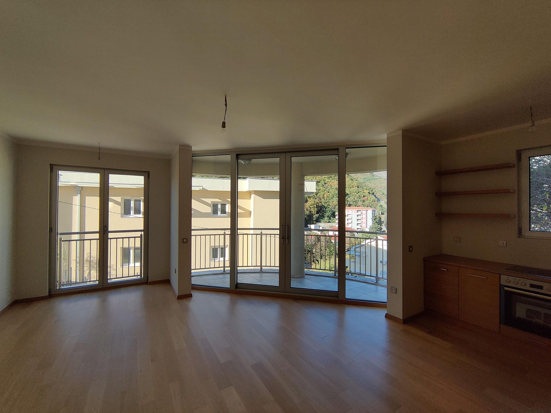Salon lumineux avec baies vitrées panoramiques et cuisine dans l’appartement, Bečići, Monténégro. Vue sur les montagnes et la terrasse, parquet au sol.
