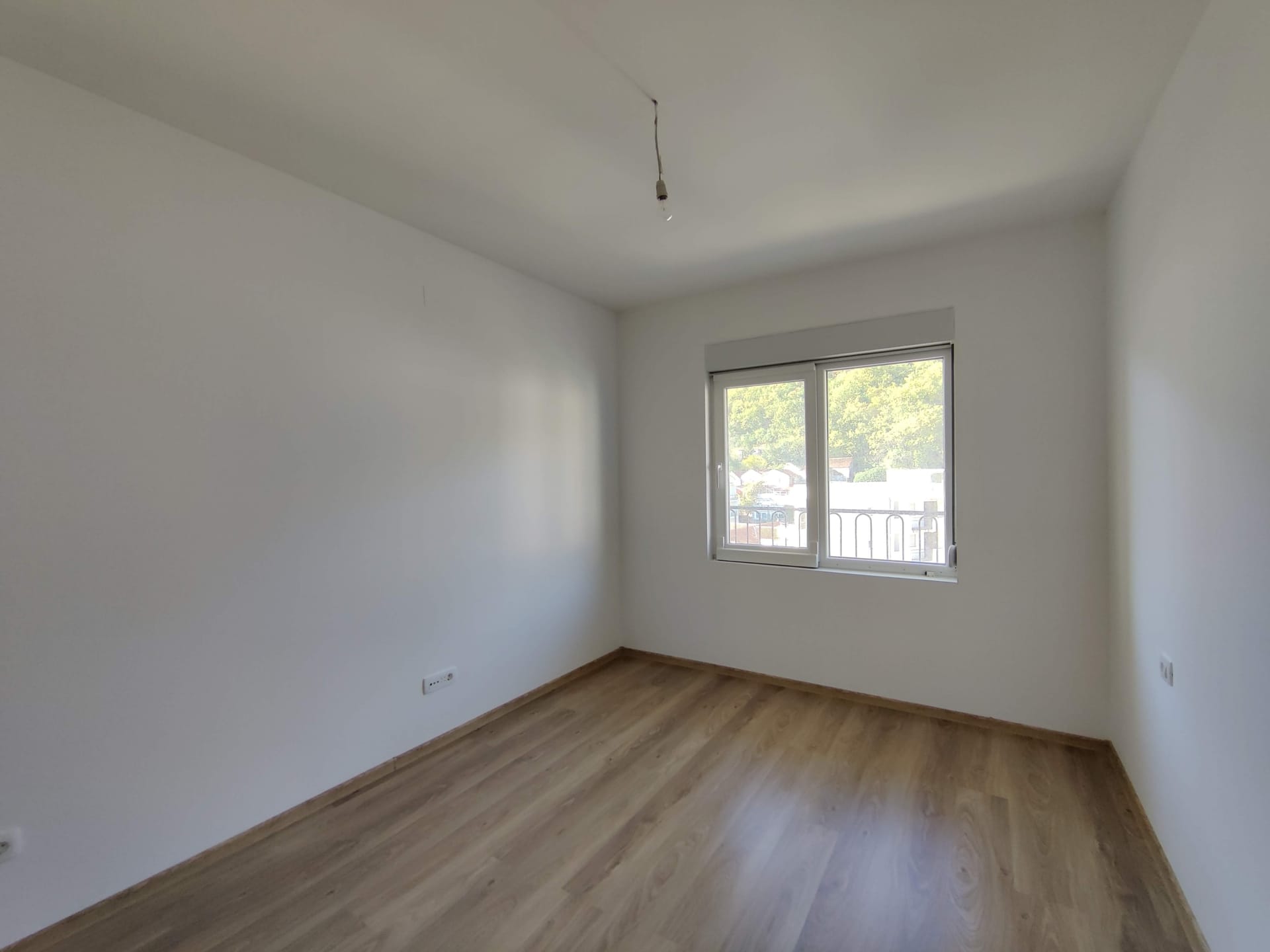 Chambre lumineuse sans meubles avec parquet stratifié et fenêtre dans un appartement à vendre à Bečići, Monténégro.