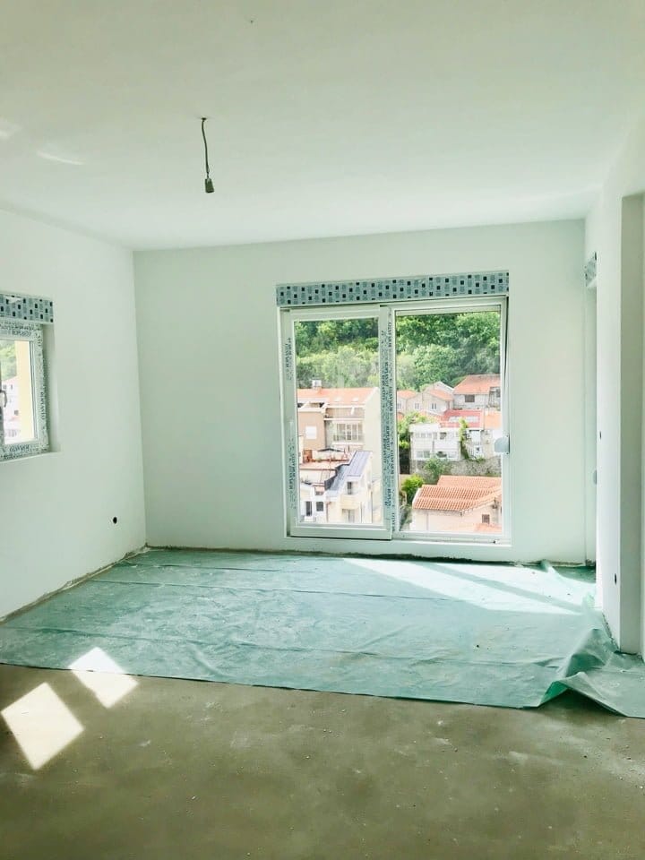 Salon lumineux non fini dans un immeuble neuf à Bečići, Monténégro, avec des fenêtres panoramiques et une vue sur la ville et les montagnes.