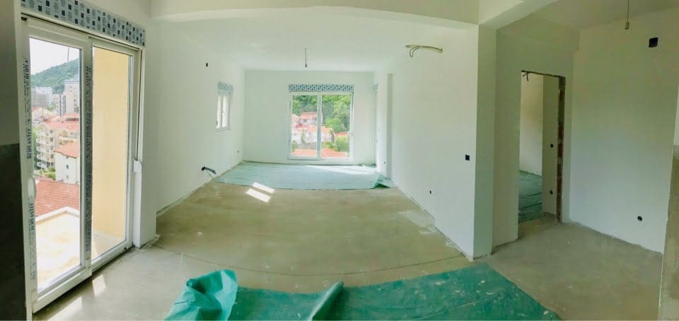 Salon spacieux non fini dans un immeuble neuf, Bečići, Monténégro : fenêtres panoramiques et vue sur les montagnes et la ville.