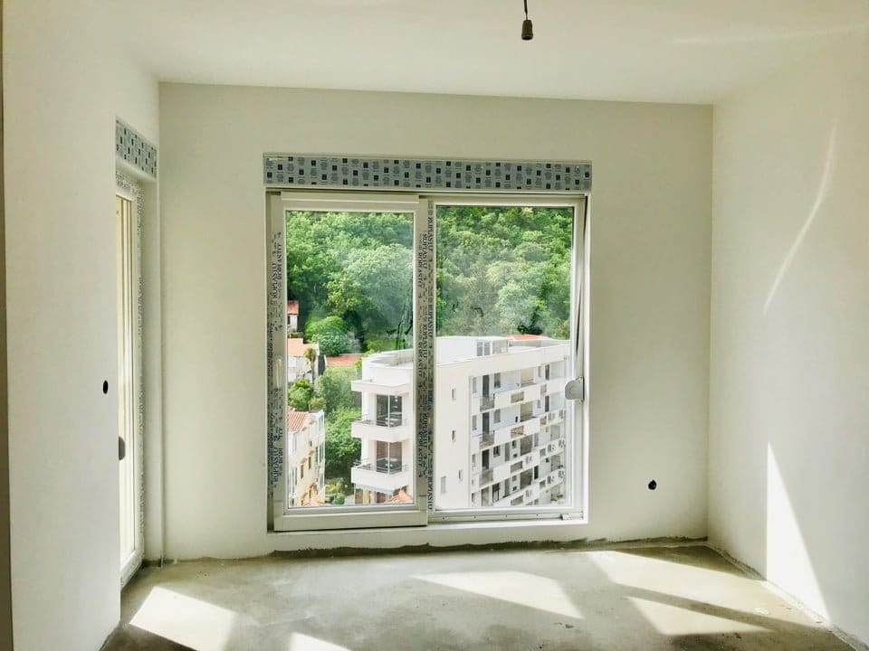 Salon dans un immeuble neuf à Bečići, Monténégro : pièce lumineuse avec une fenêtre panoramique et vue sur les montagnes et la ville.