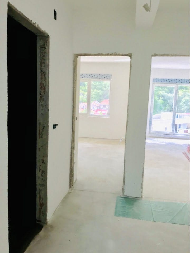 Salon dans un appartement non fini à Bečići, Monténégro : murs clairs, ouvertures de porte et vue sur la verdure depuis les fenêtres.