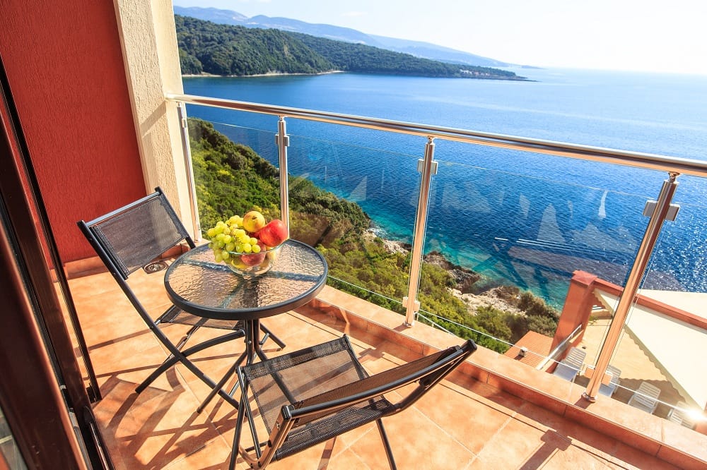 Balkon mit Möbeln und Meerblick in einer Wohnanlage, Dobre Vode, Montenegro. Auf dem Tisch Früchte, Glasgeländer.