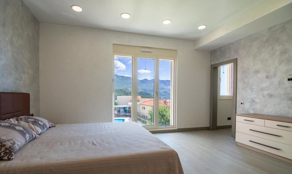 Karadağ, Bečići’deki villada modern yatak odası: çift kişilik yatak, şifonyer ve pencereden panoramik dağ manzarası.