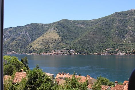 Panoramablick auf die Bucht von Kotor und die Berge aus dem Fenster der Wohnung in Dobrota, Montenegro, mit Ziegeldächern im Vordergrund.