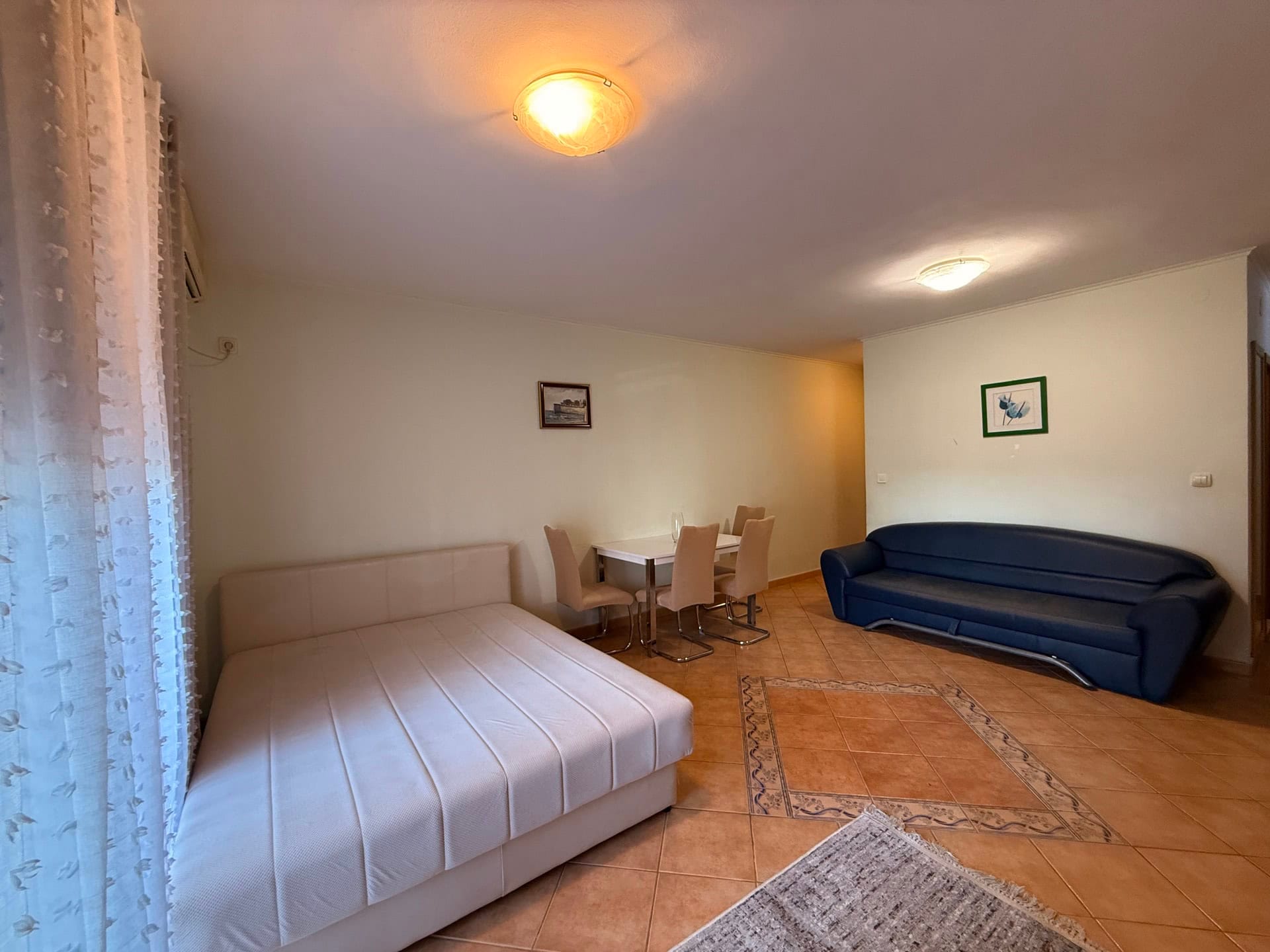 Living room in an apartment in Budva, Montenegro: double bed, blue sofa, dining area, and tiled floor.