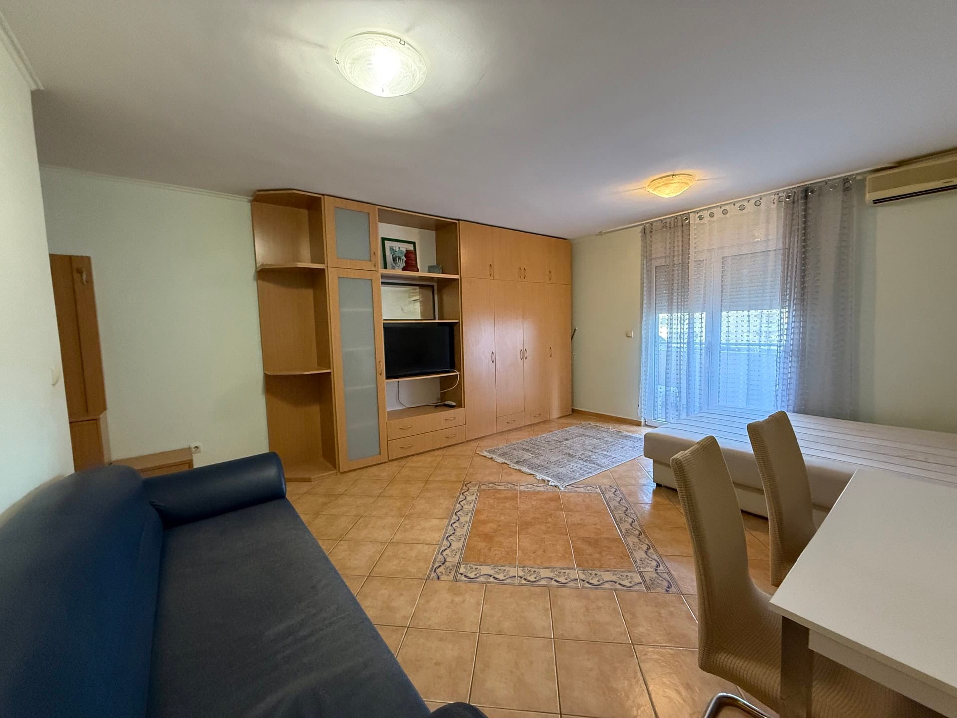 Living room in an apartment in Budva, Montenegro: blue sofa, wooden wall unit, dining area, and access to the balcony.