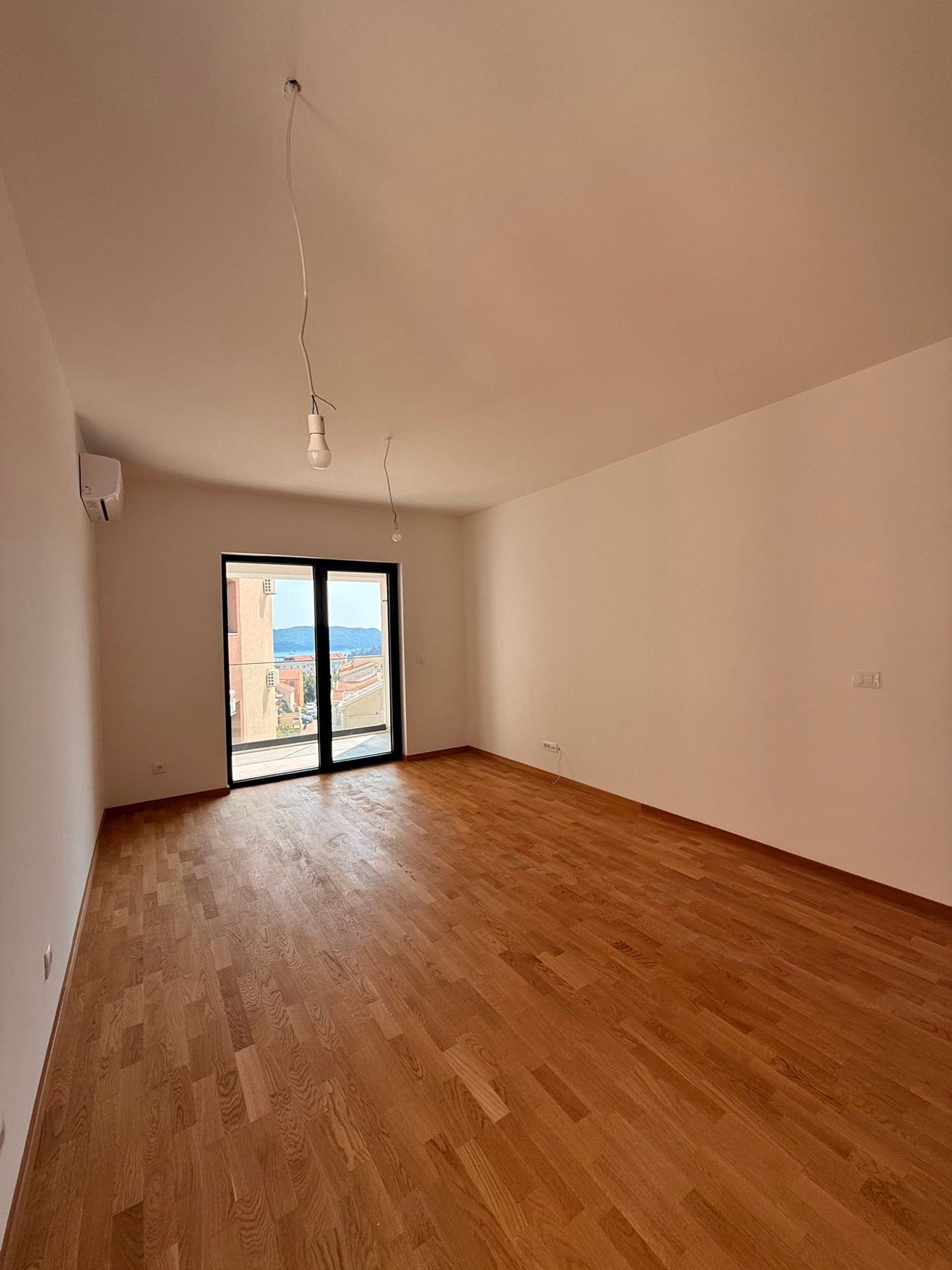 Svetao dnevni boravak sa parketom i izlazom na balkon u novom stanu u Bečićima, Crna Gora, pogled na more i planine.