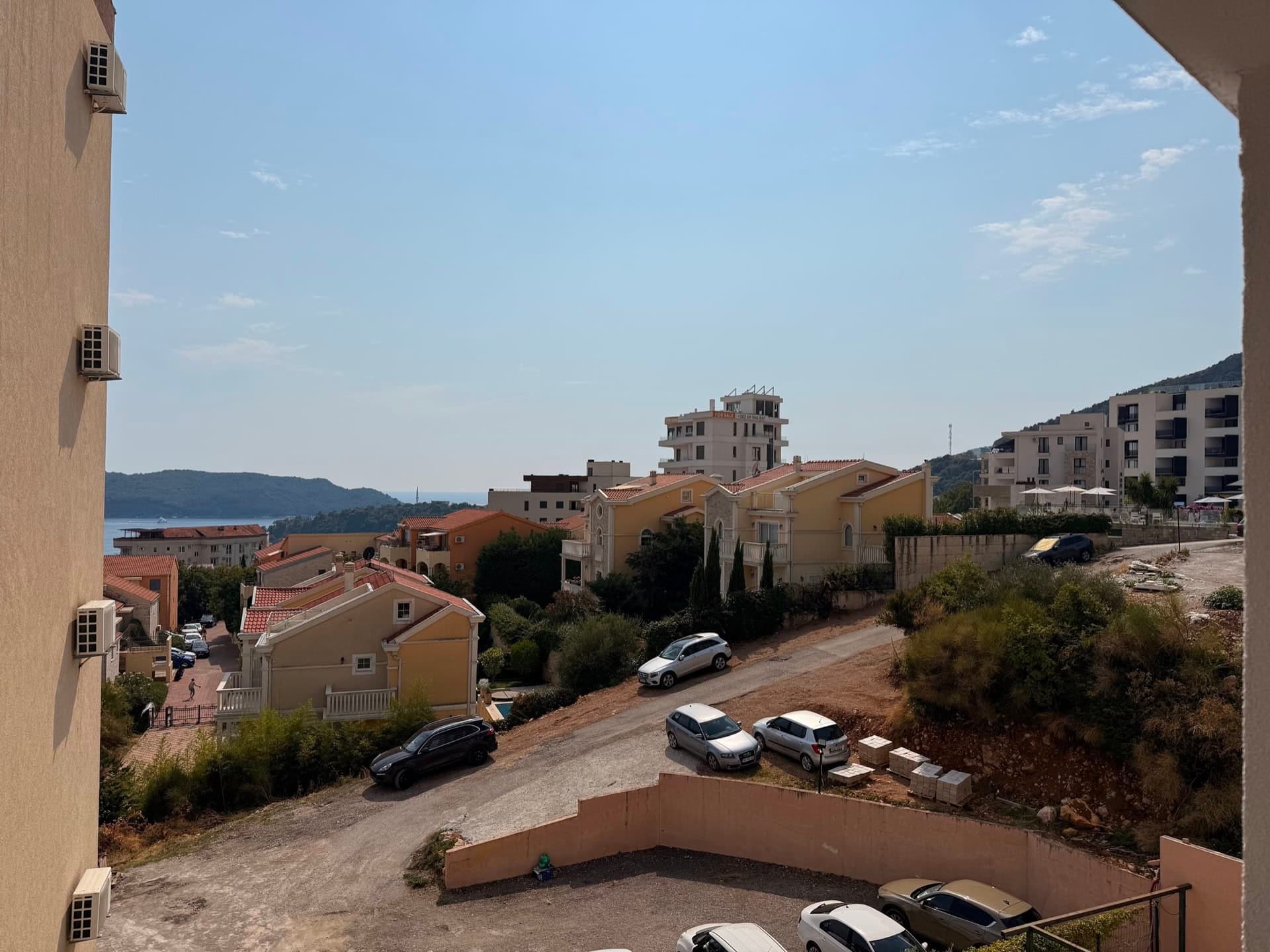 Pogled sa balkona stana u Bečićima, Crna Gora: panorama stambenog kompleksa, planina i morske obale po sunčanom danu.
