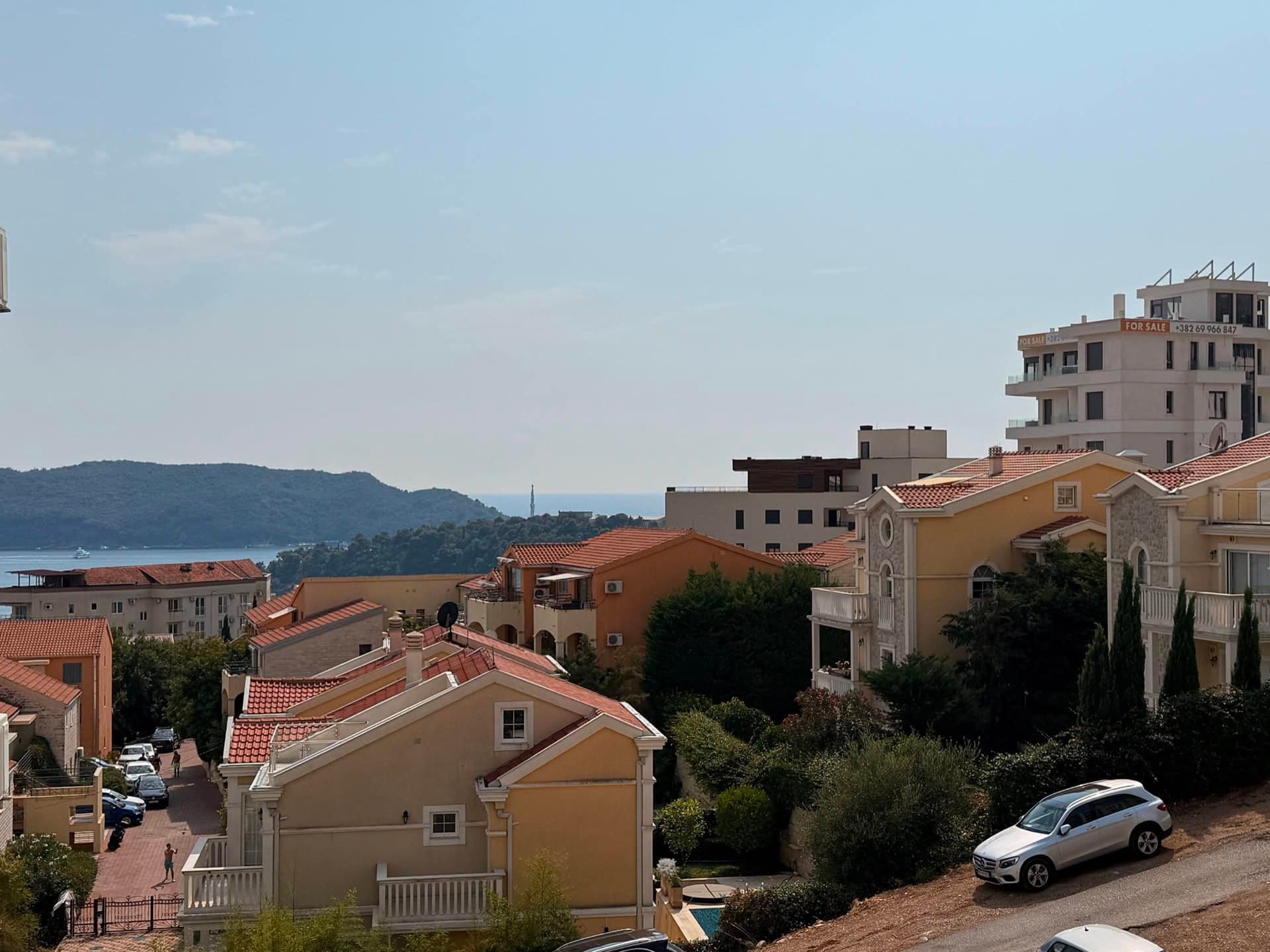 Panoramski pogled na more i stambeni kompleks u Bečićima, Crna Gora: prijatne kuće sa crepnim krovovima i zeleni brežuljci.