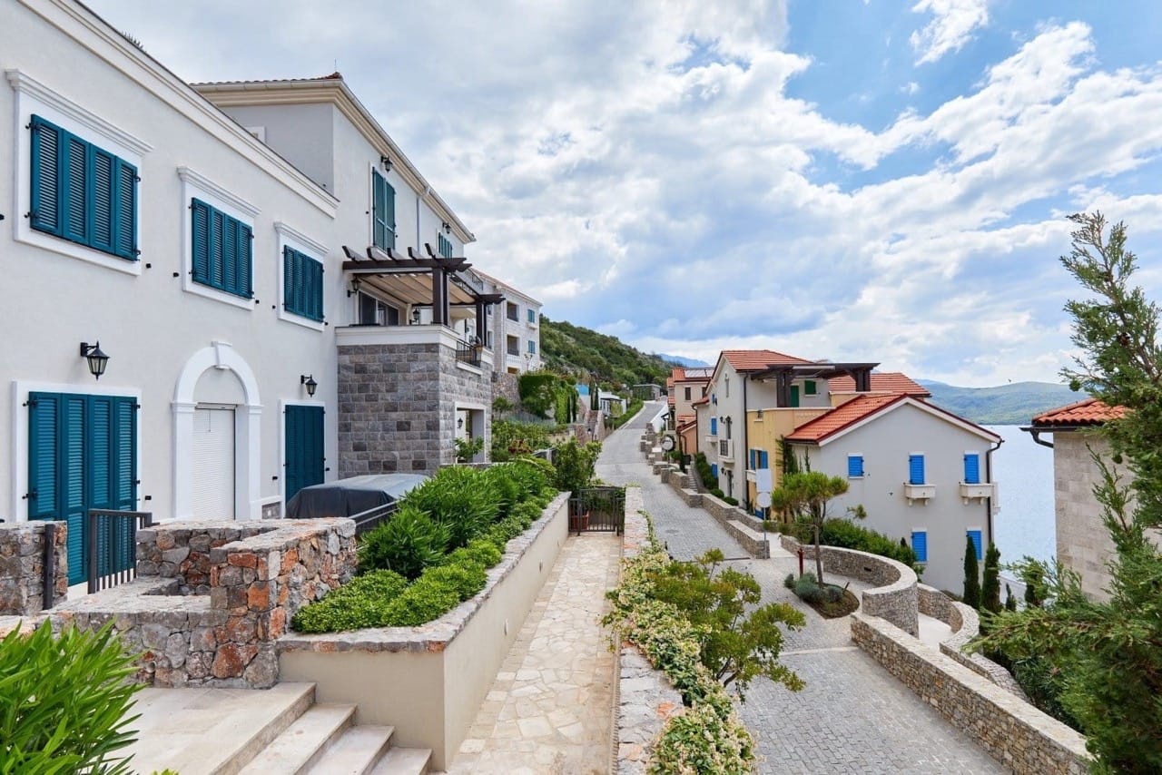Straße der exklusiven Wohnanlage Lustica Bay in Radovici, Montenegro: Steinhäuser mit blauen Fensterläden und Meerblick.