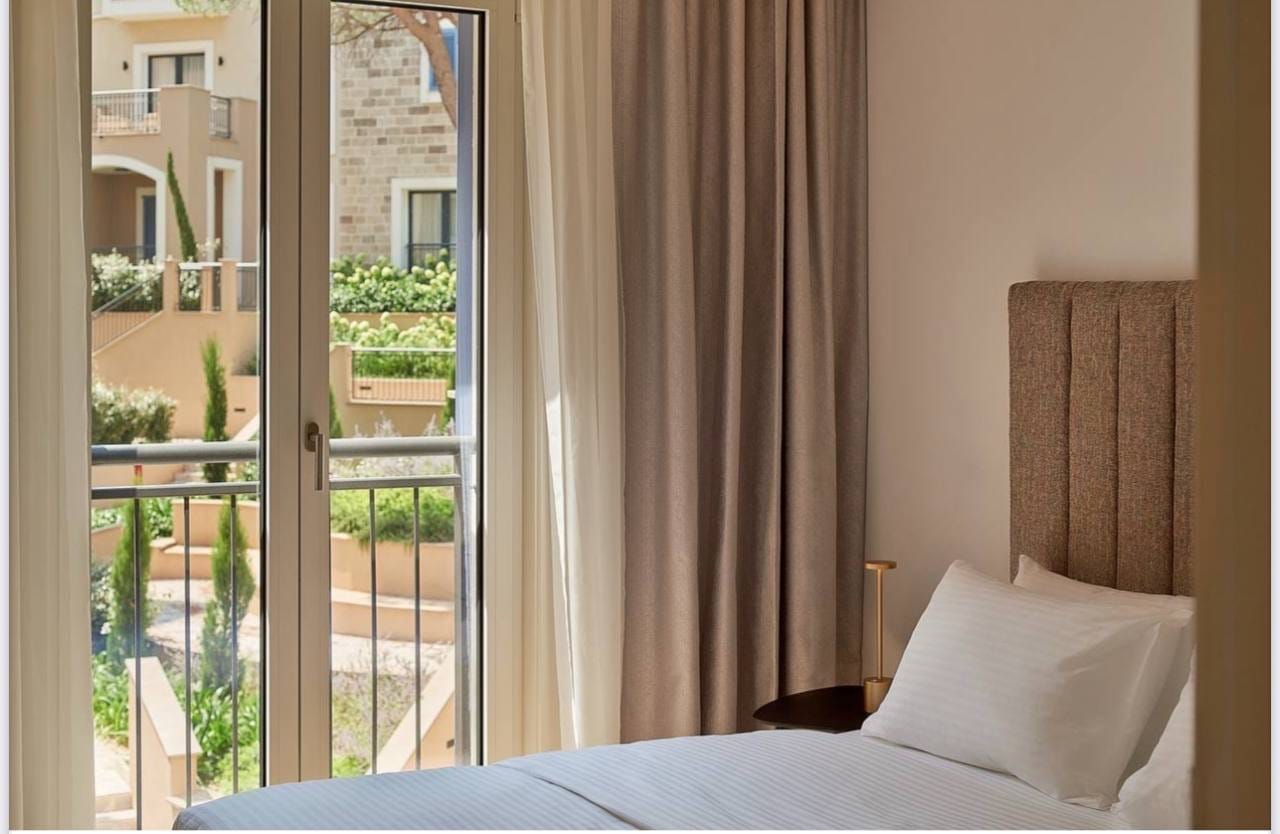 Spavaća soba u apartmanima Luštica Bay, Crna Gora: moderan krevet sa mekanim uzglavljem i pogled na terasu kompleksa.