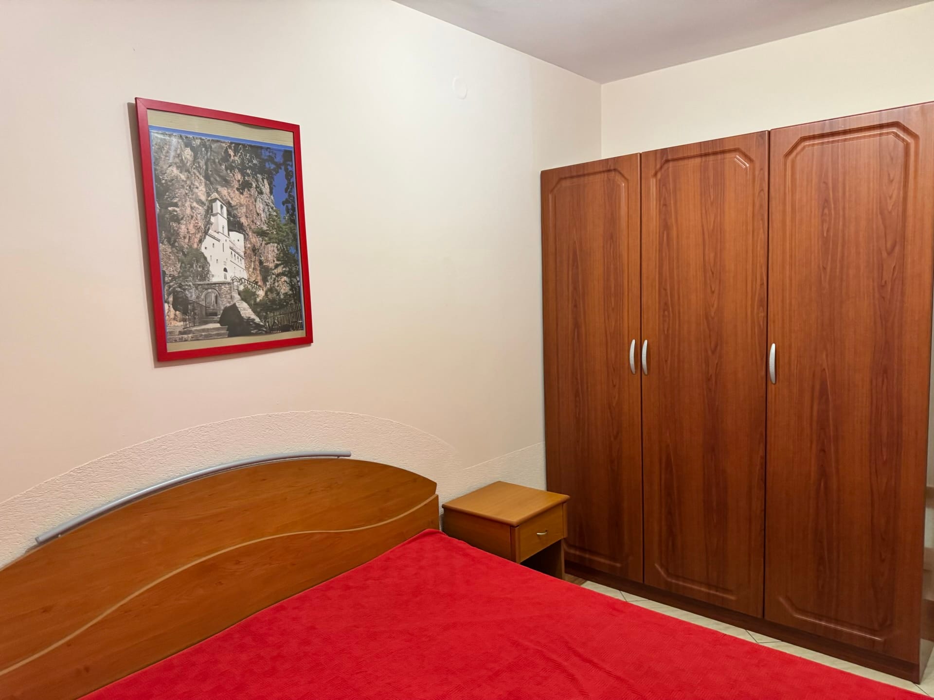 Spavaća soba u stanu u Budvi, Crna Gora: bračni krevet, drveni ormar i slika manastira Ostrog.