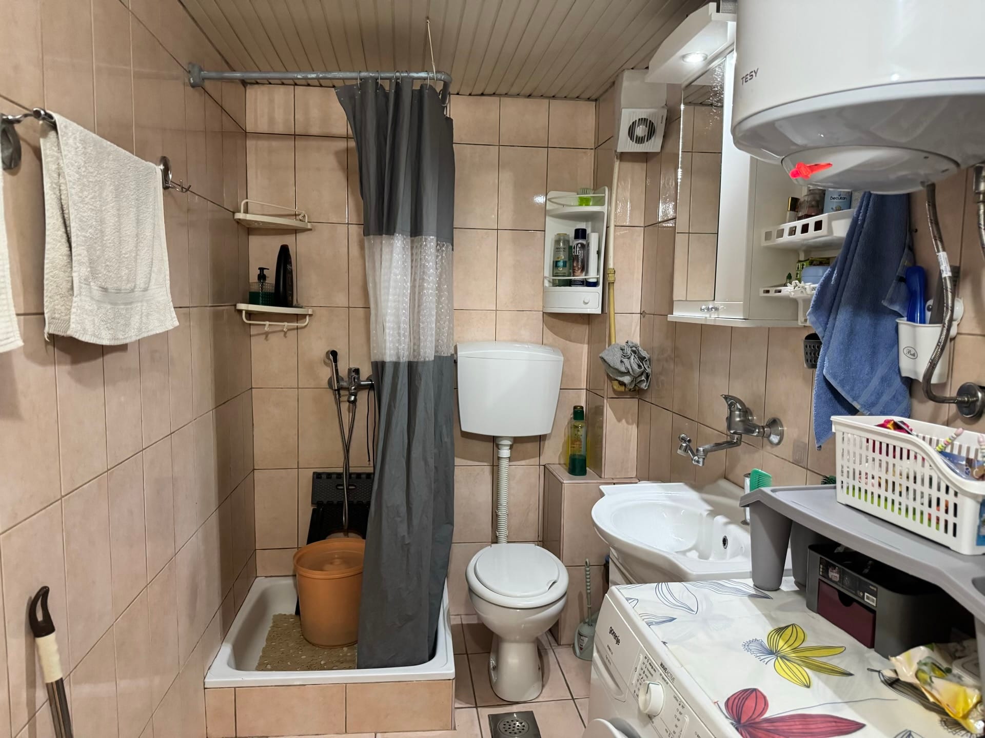 Kupatilo u stanu u Bečićima, Crna Gora: pločice, tuš zona sa zavesom, WC šolja i veš mašina.