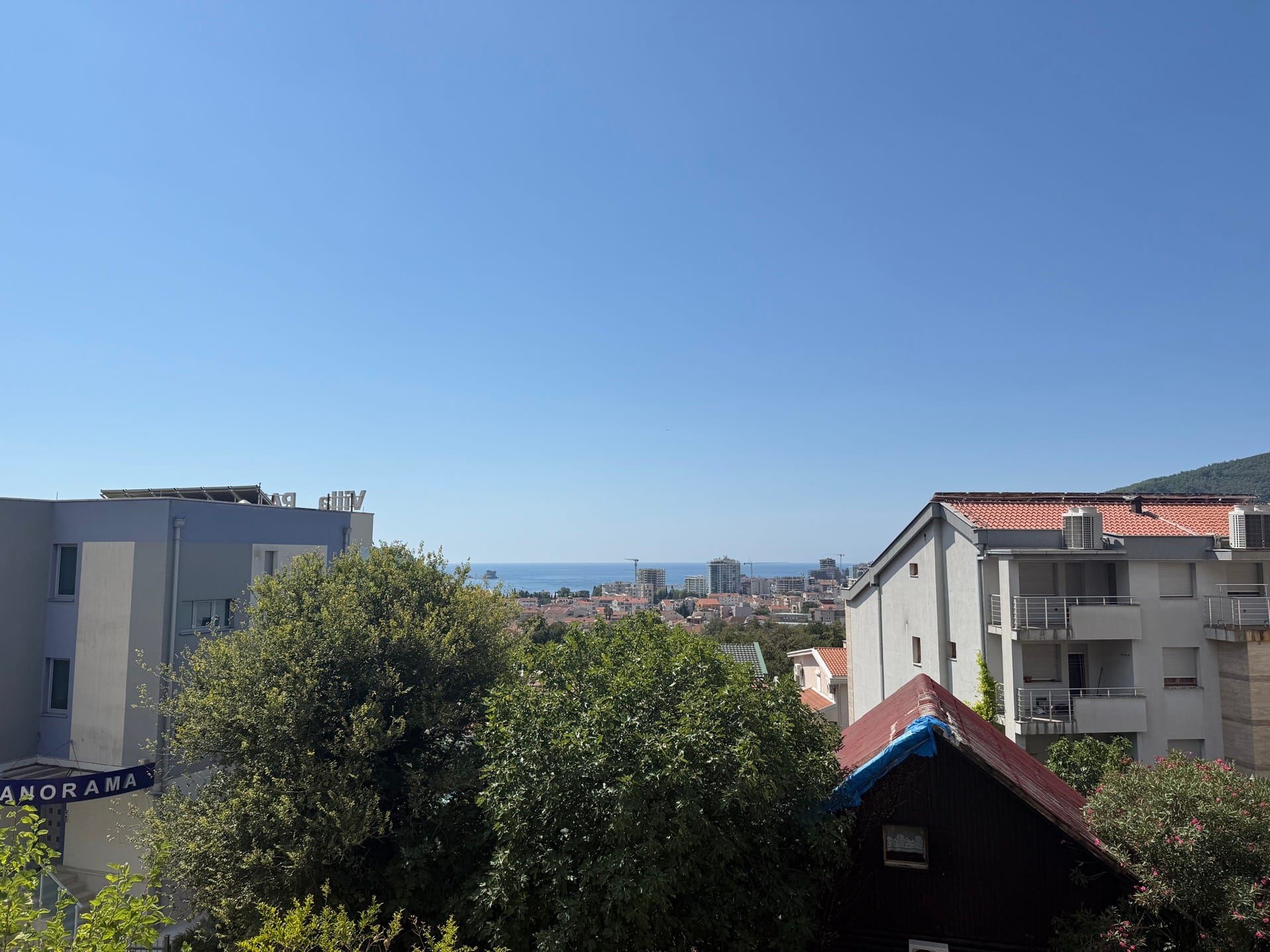 Panoramski pogled na more i grad Budvu, Crna Gora, iz prozora stana sa zelenim drvećem u prvom planu.