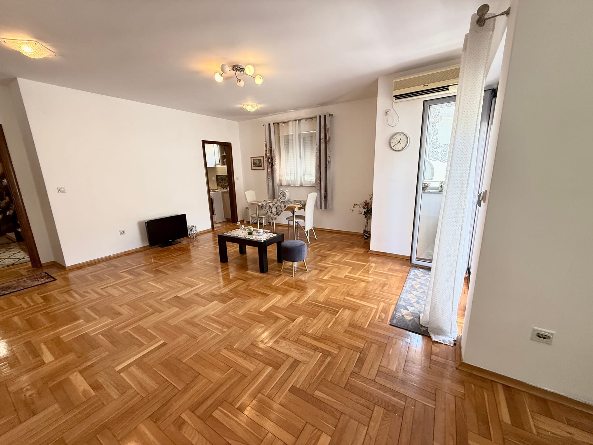 Prostrana dnevna soba sa parketom i izlazom na balkon u stanu, Budva, Crna Gora. Namešteni deo za odmor i trpezarija.