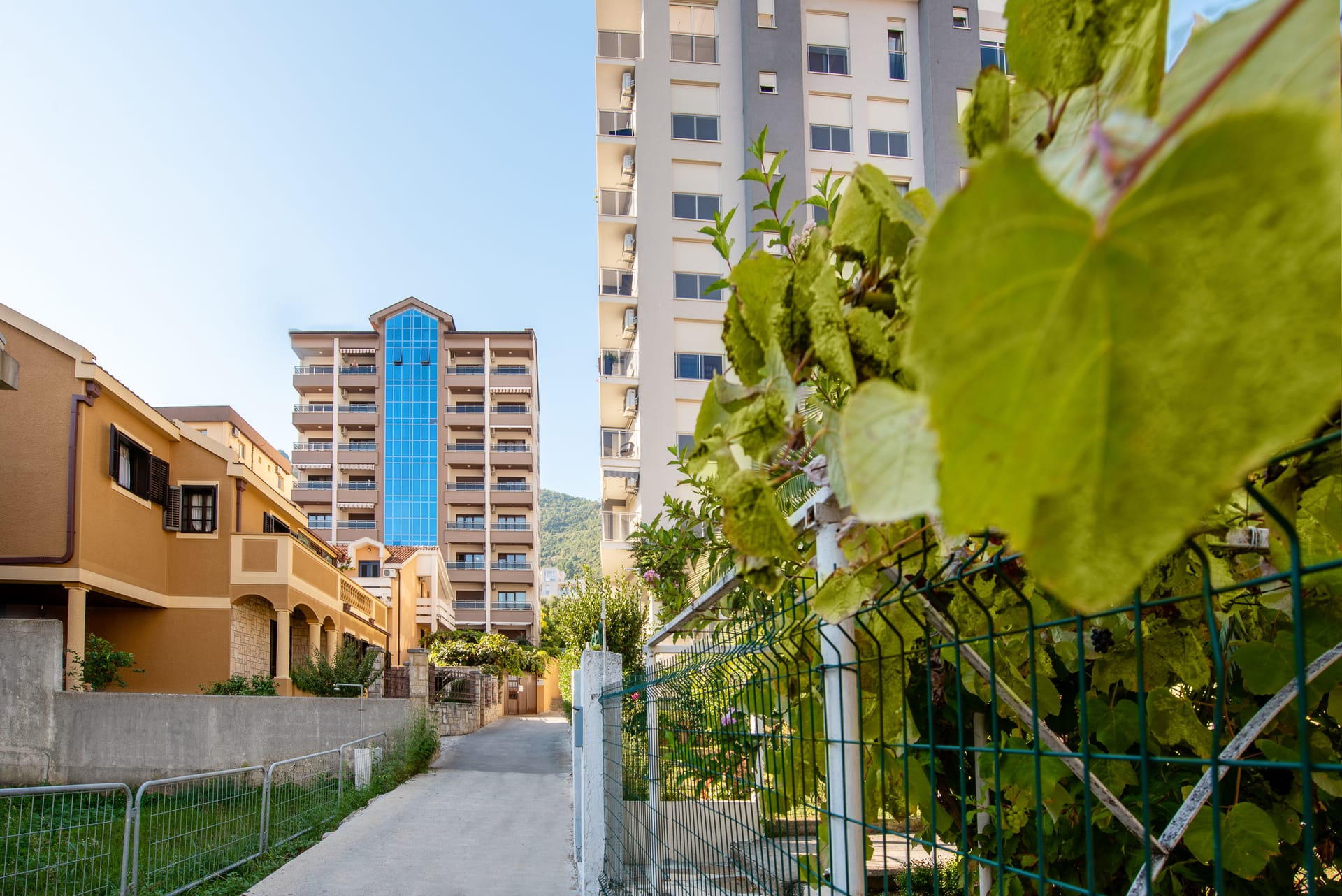 Pogled na moderne višespratne stambene zgrade u Budvi, Crna Gora, sa uskom ulicom i zelenom ogradom u prvom planu.