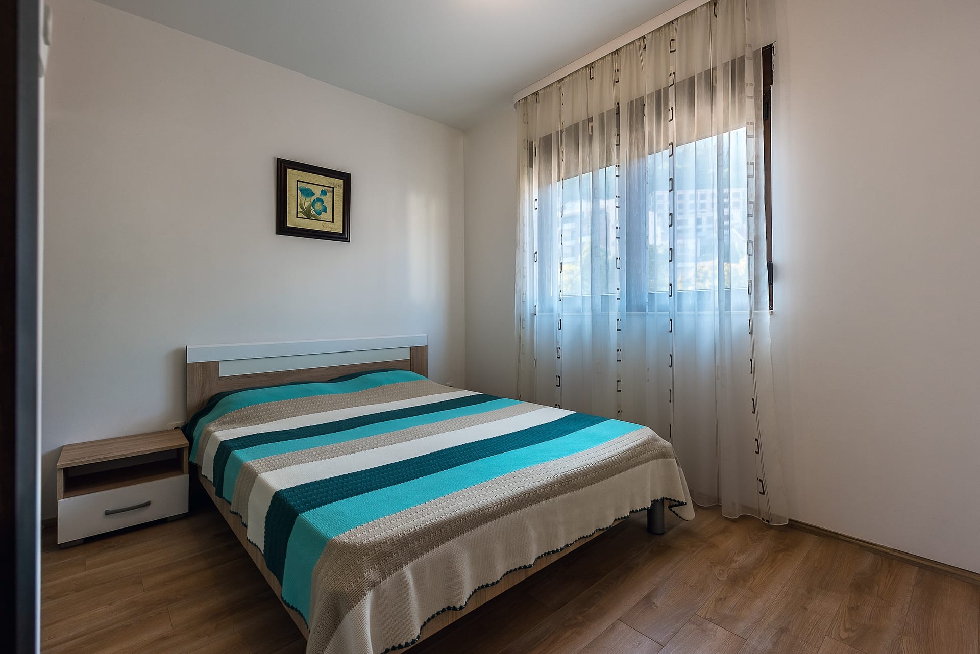 Svetla spavaća soba sa bračnim krevetom u apartmanima Budve, Crna Gora: laminat, noćni ormarić i prozor sa tilom.