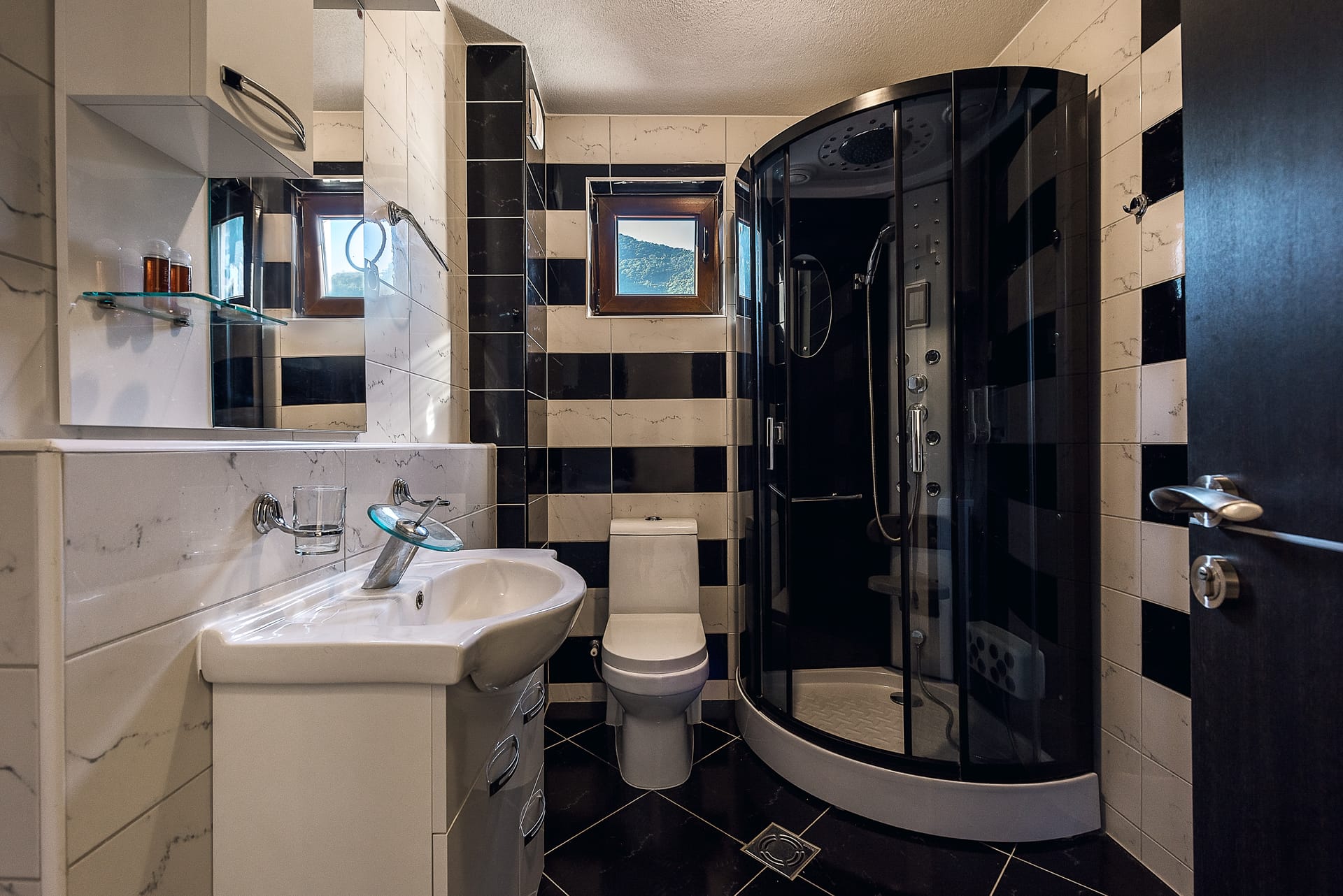 Moderno kupatilo sa tuš kabinom u apartmanima Budve, Crna Gora: crno-bele pločice i pogled na planine.