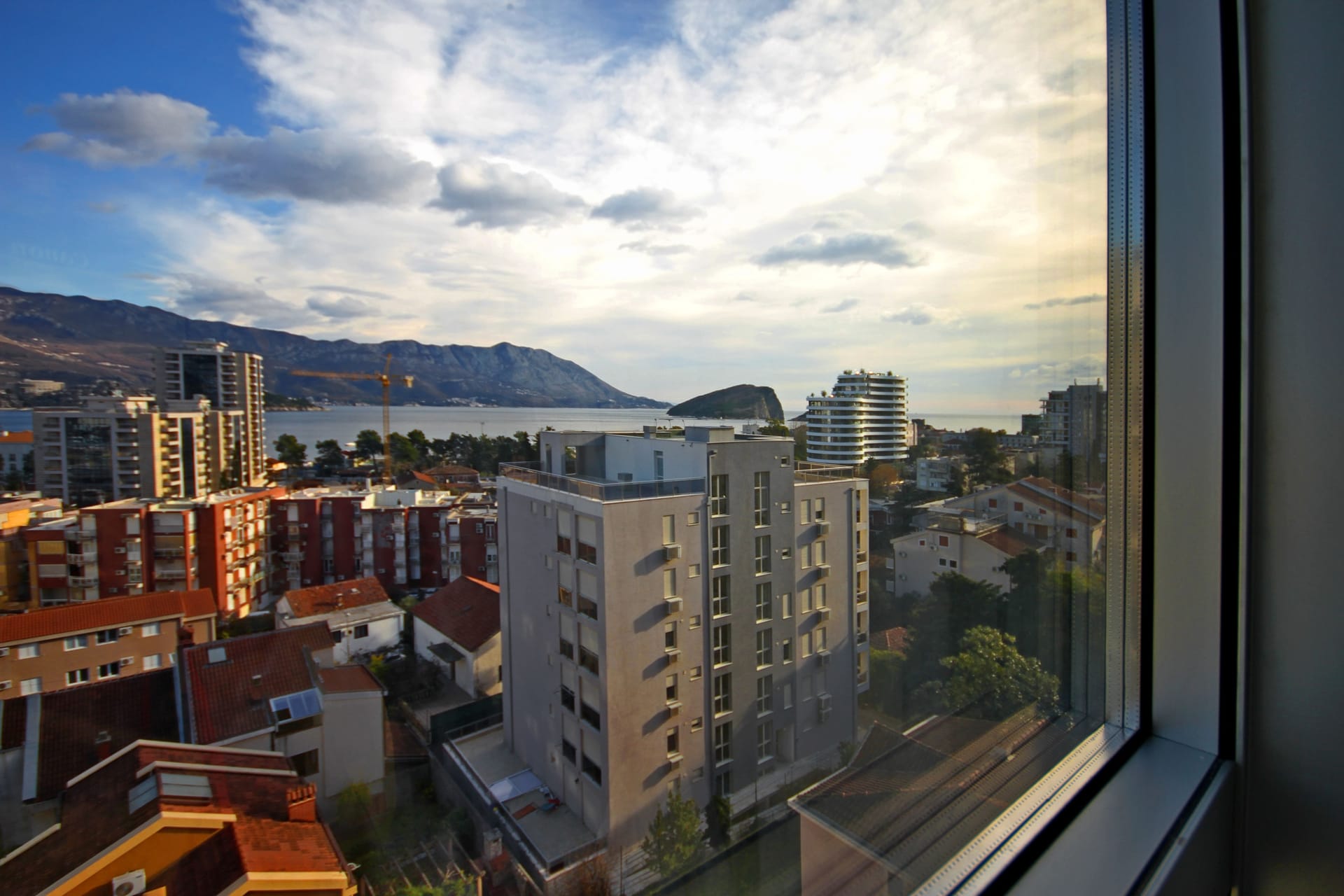 Pogled sa prozora stana u Budvi, Crna Gora: panorama grada, moderne stambene zgrade, more i ostrvo Svetog Nikole.