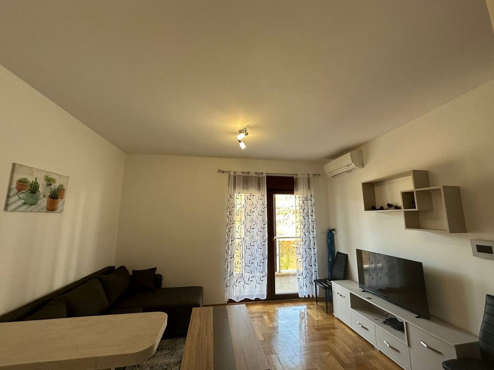 Svetla dnevna soba u apartmanima, Bečići, Crna Gora: sofa, TV zona, parket i izlaz na balkon.