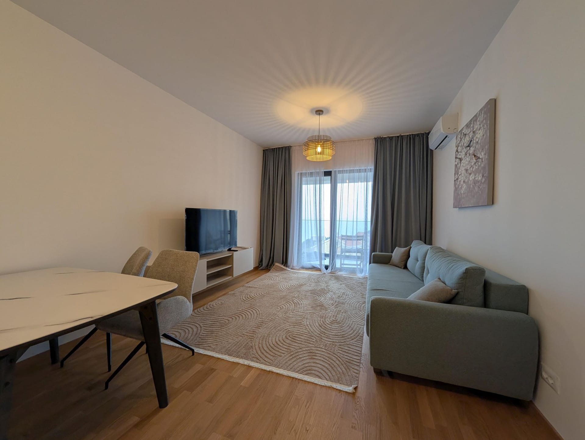 Moderna dnevna soba u apartmanima Bečići, Crna Gora: sofa, trpezarijski deo, TV i izlaz na terasu sa pogledom na more.