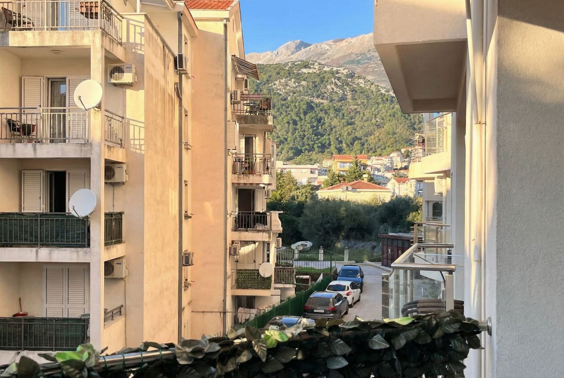 Pogled sa balkona stana u Budvi, Crna Gora: stambene zgrade, parking i panoramski pejzaž zelenih planina.
