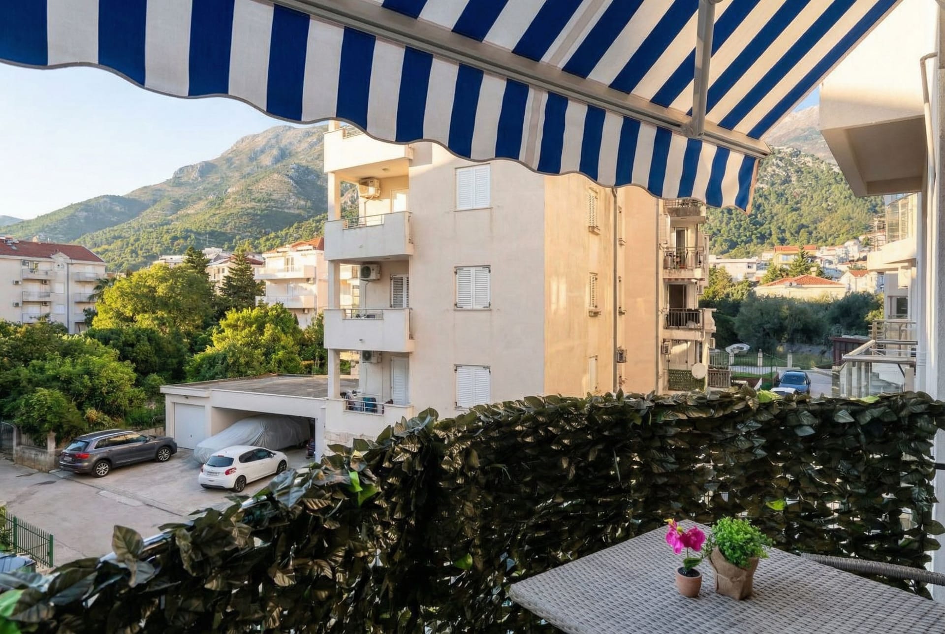 Pogled sa balkona stana u Budvi, Crna Gora: prugasta tenda, pleteni stočić i panorama planina i grada.