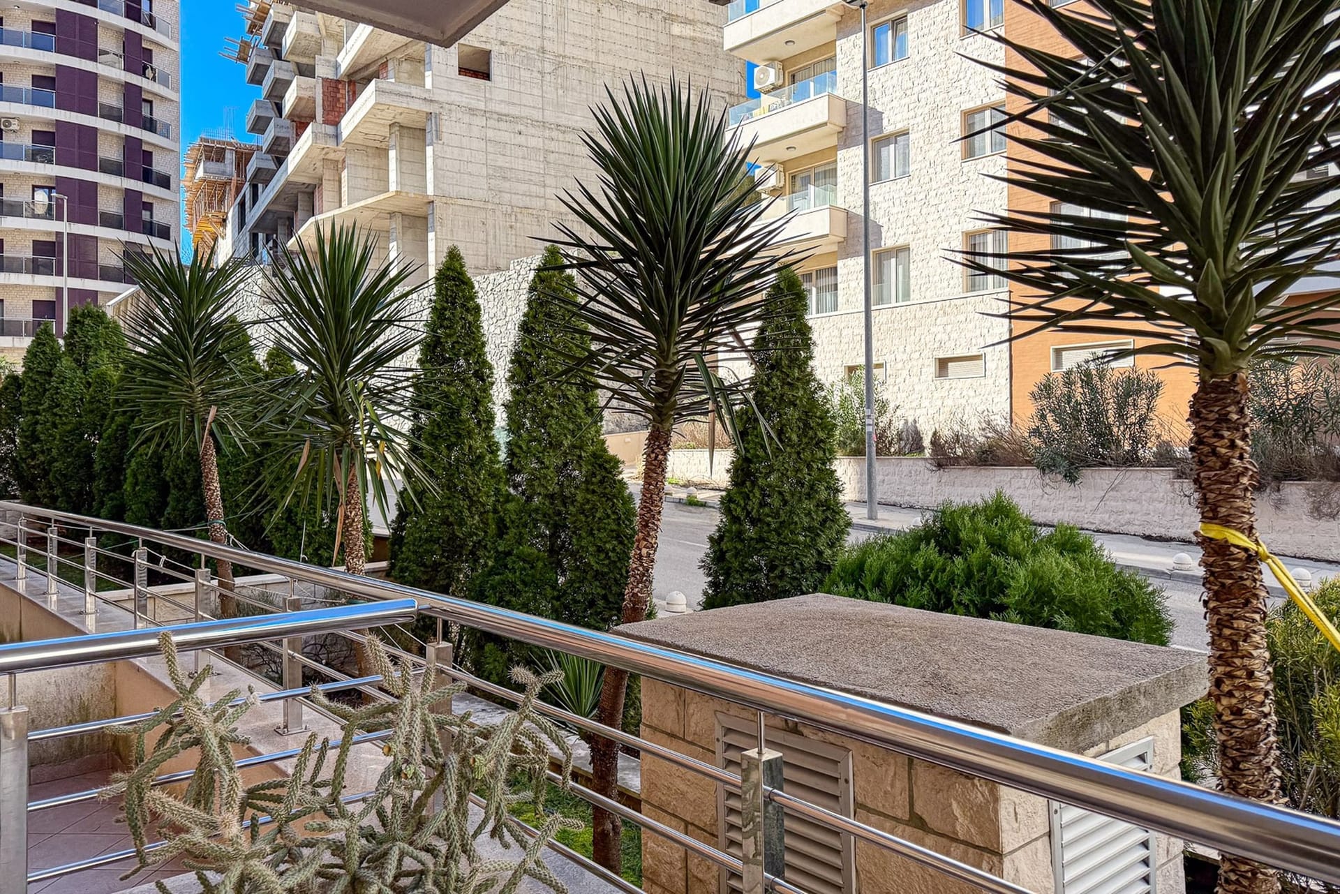 Pogled sa balkona stana u Bečićima, Crna Gora: palme, tuje i moderne stambene zgrade na pozadini plavog neba.