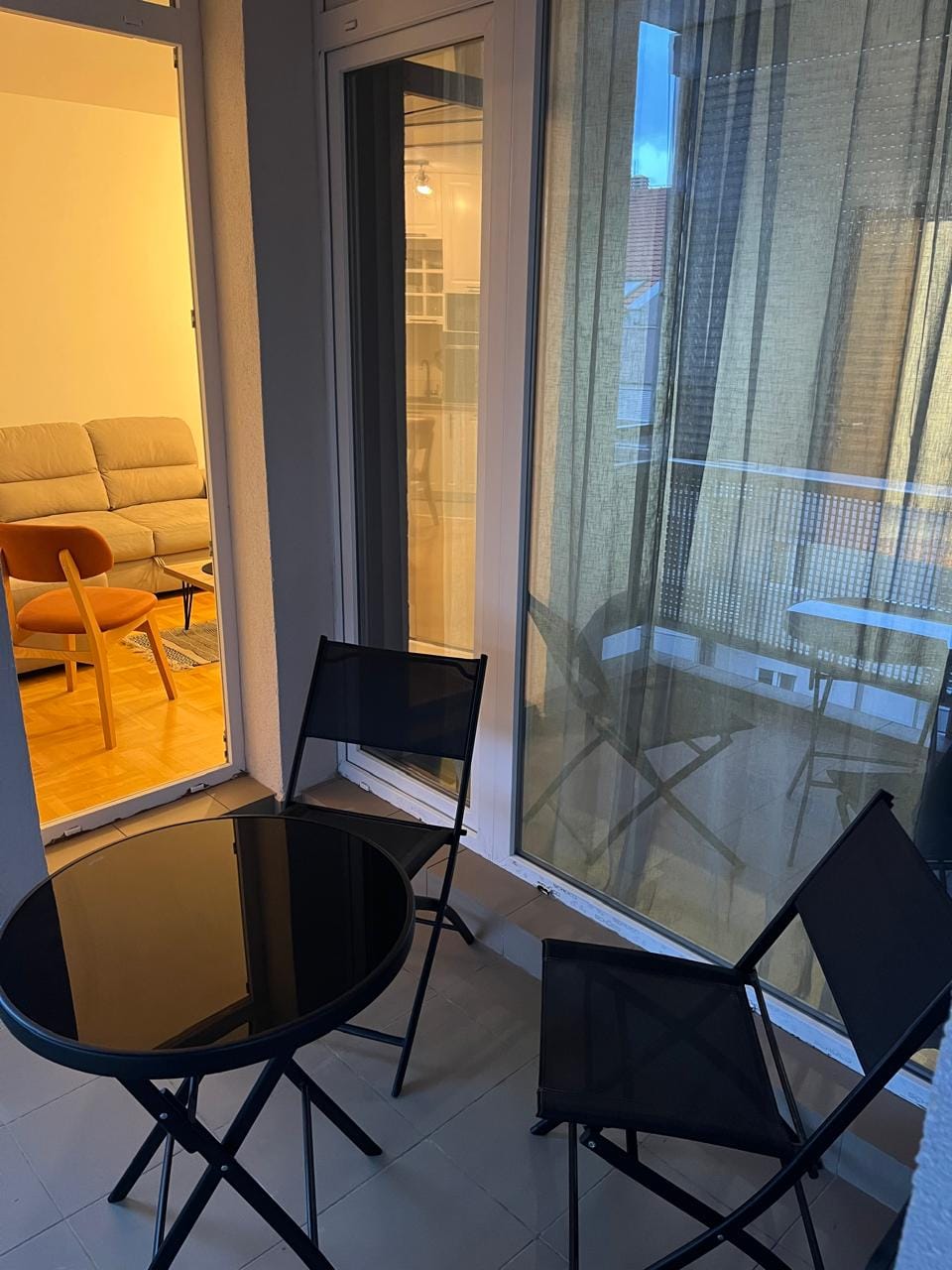 Balkon sa crnim stolom i stolicama u stanu u Budvi, Crna Gora, sa pogledom na prijatnu dnevnu sobu sa mekanom sofom.
