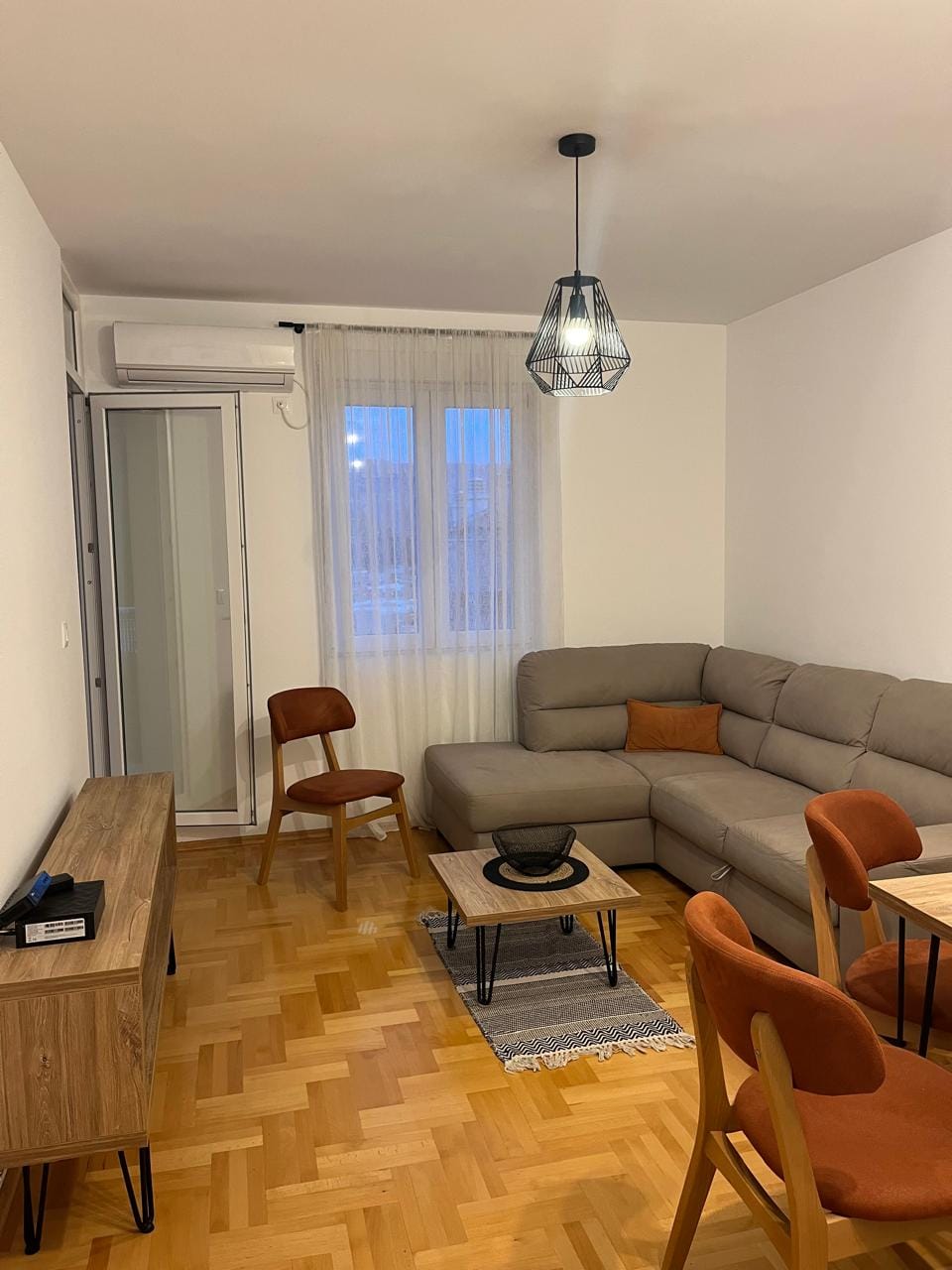 Ugodna dnevna soba sa sivim ugaonim sofom i parketom u stanu, Budva, Crna Gora. Moderan enterijer sa izlazom na balkon.
