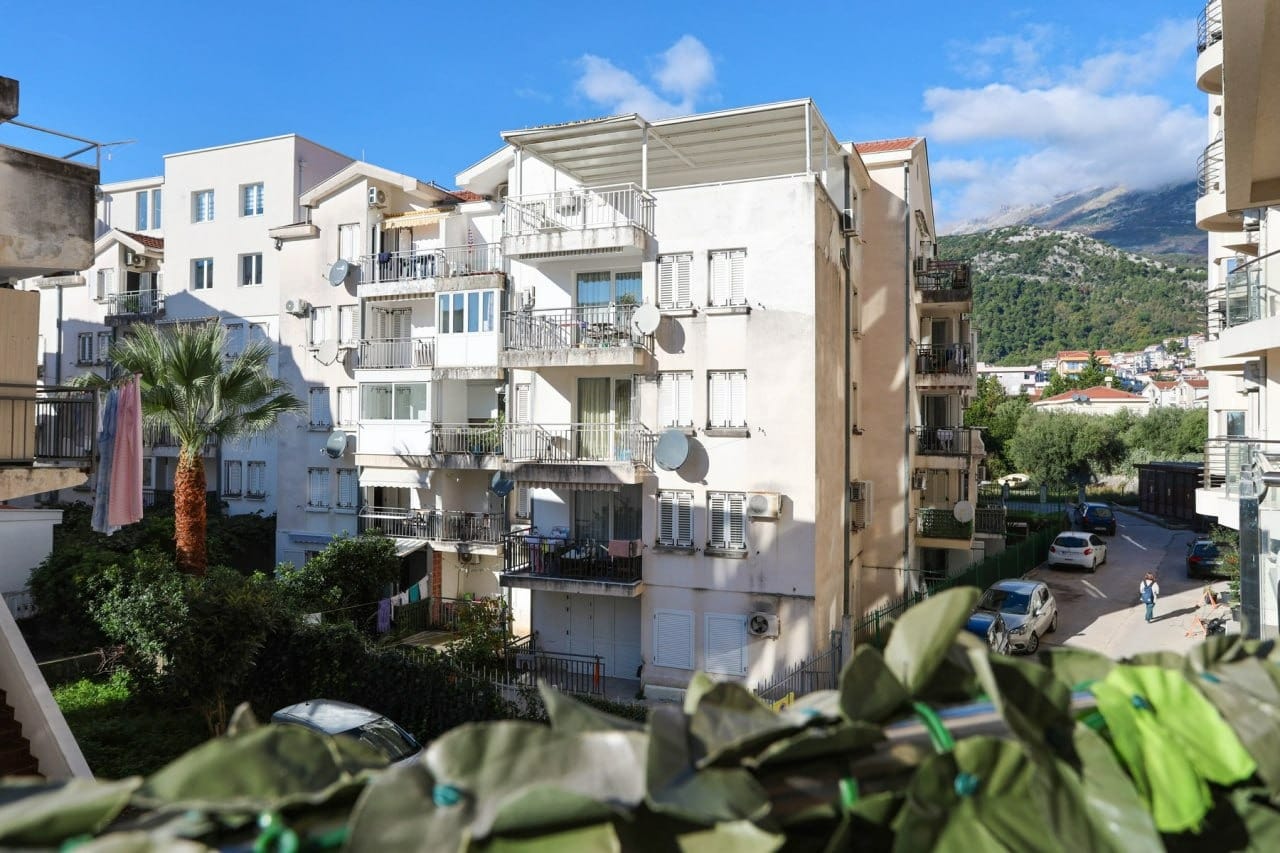 Pogled na stambenu zgradu sa balkonima i palmom u gradu Budvi, Crna Gora. Fasada zgrade sa terasama i planinski pejzaž u pozadini.