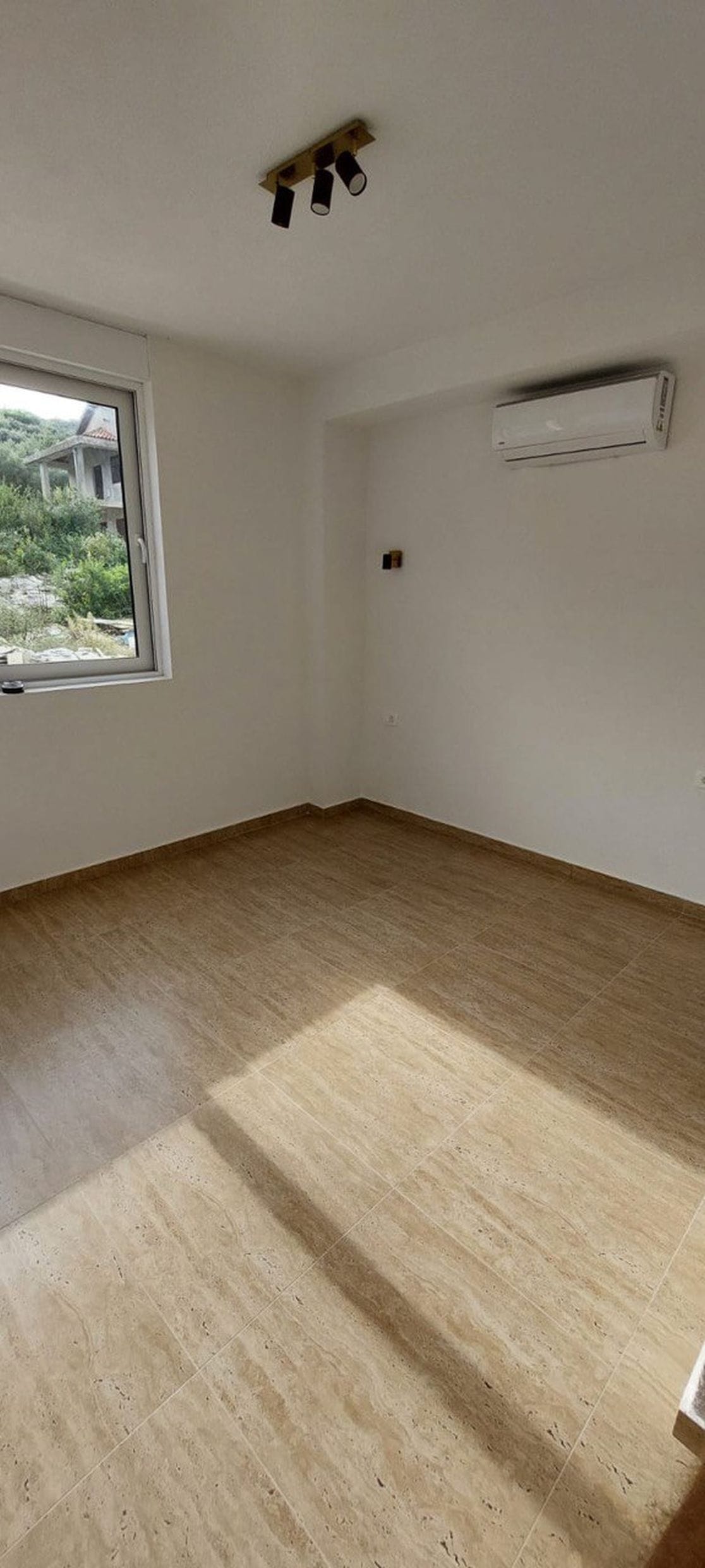Osvetljena dnevna soba u taunhausu, Luštica, Crna Gora: pločice na podu, klima-uređaj i pogled kroz prozor na prirodu.