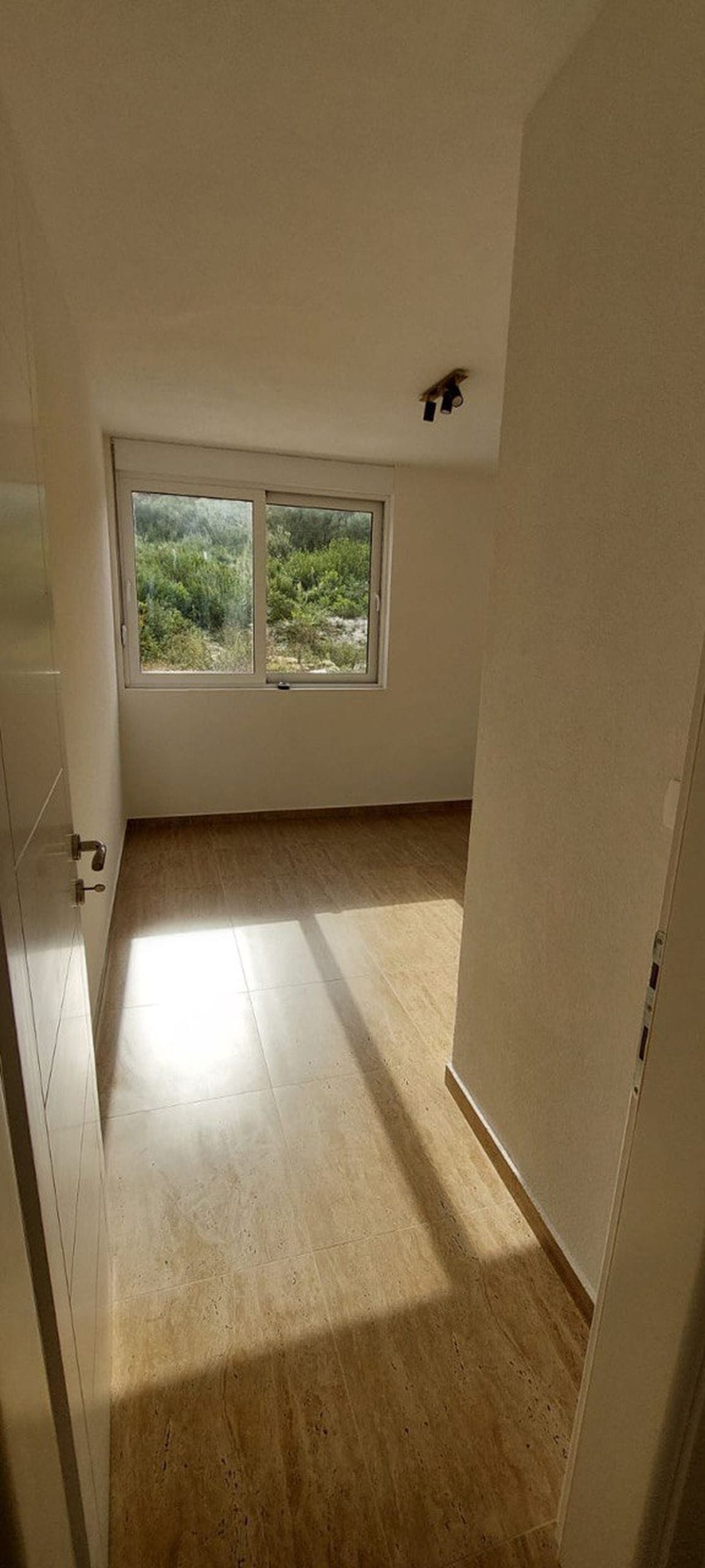 Svetla spavaća soba u taunhausu, Luštica, Crna Gora: pločice na podu, beli zidovi i pogled na zelenilo kroz prozor.