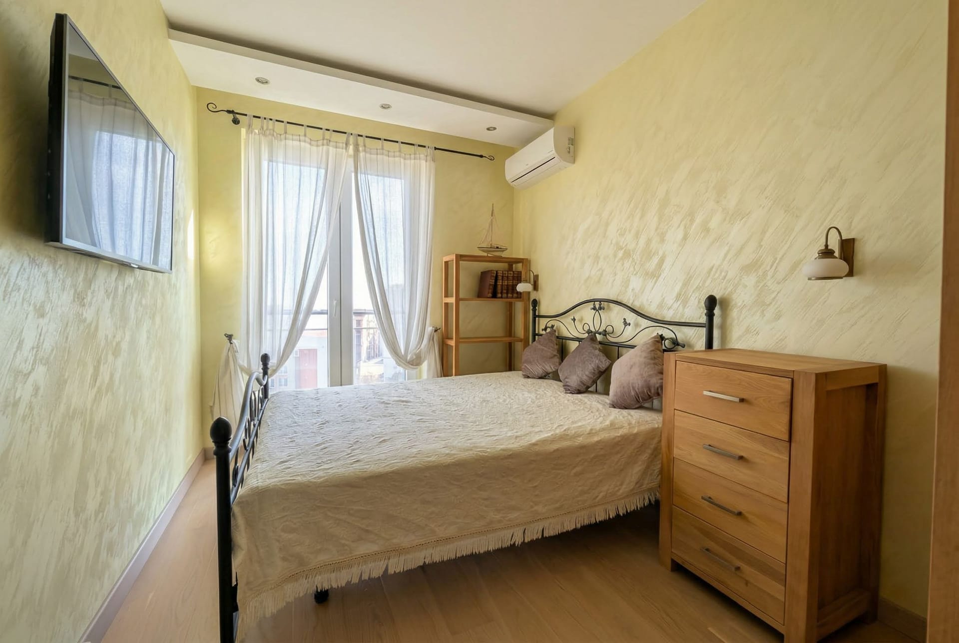 Ugodna spavaća soba sa kovanim krevetom i drvenim komodom u stanu, Bečići, Crna Gora. Svetli zidovi i izlaz na balkon.