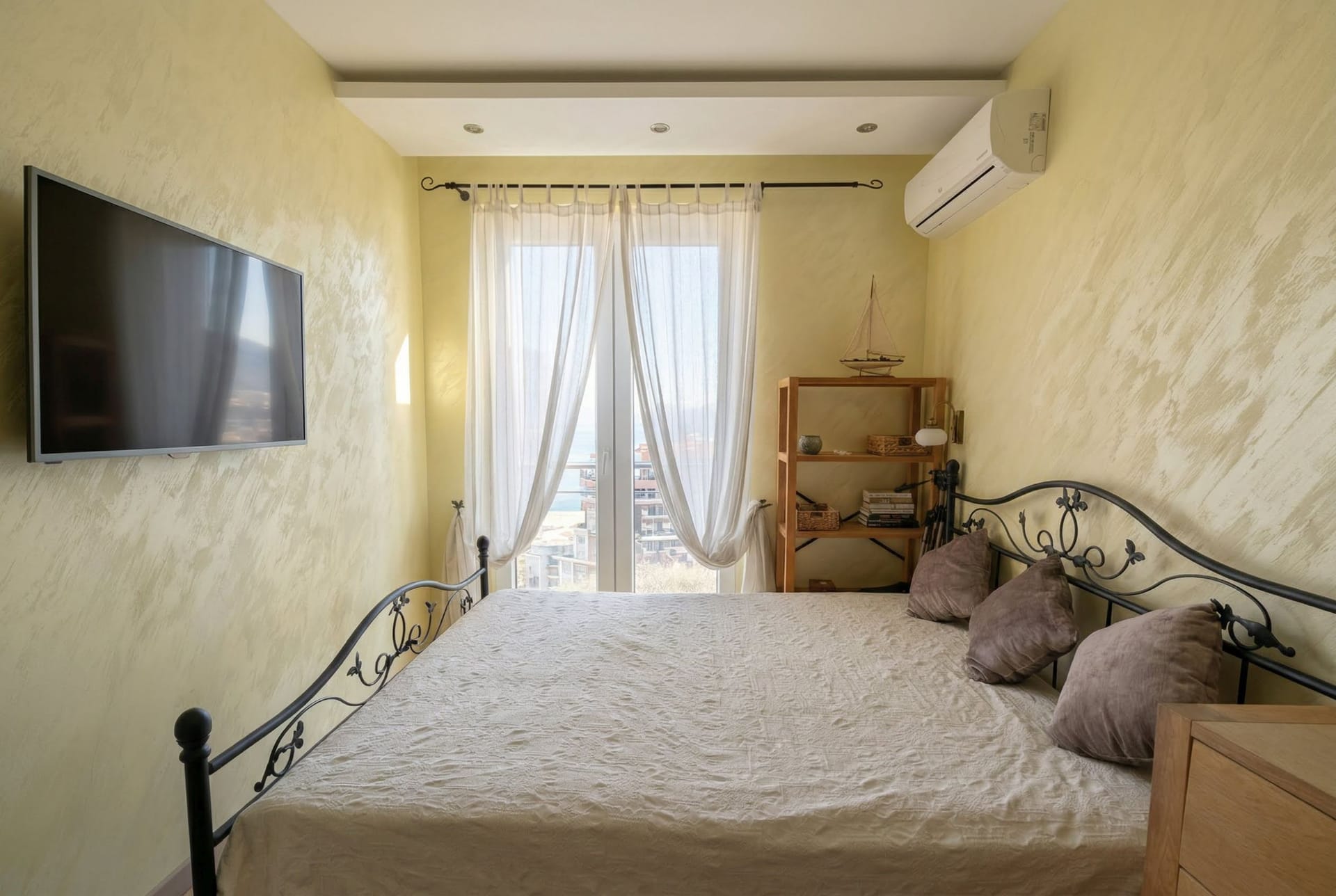 Spavaća soba sa kovanim krevetom i pogledom na more u stanu u Bečićima, Crna Gora. Svetli zidovi, TV i izlaz na balkon.