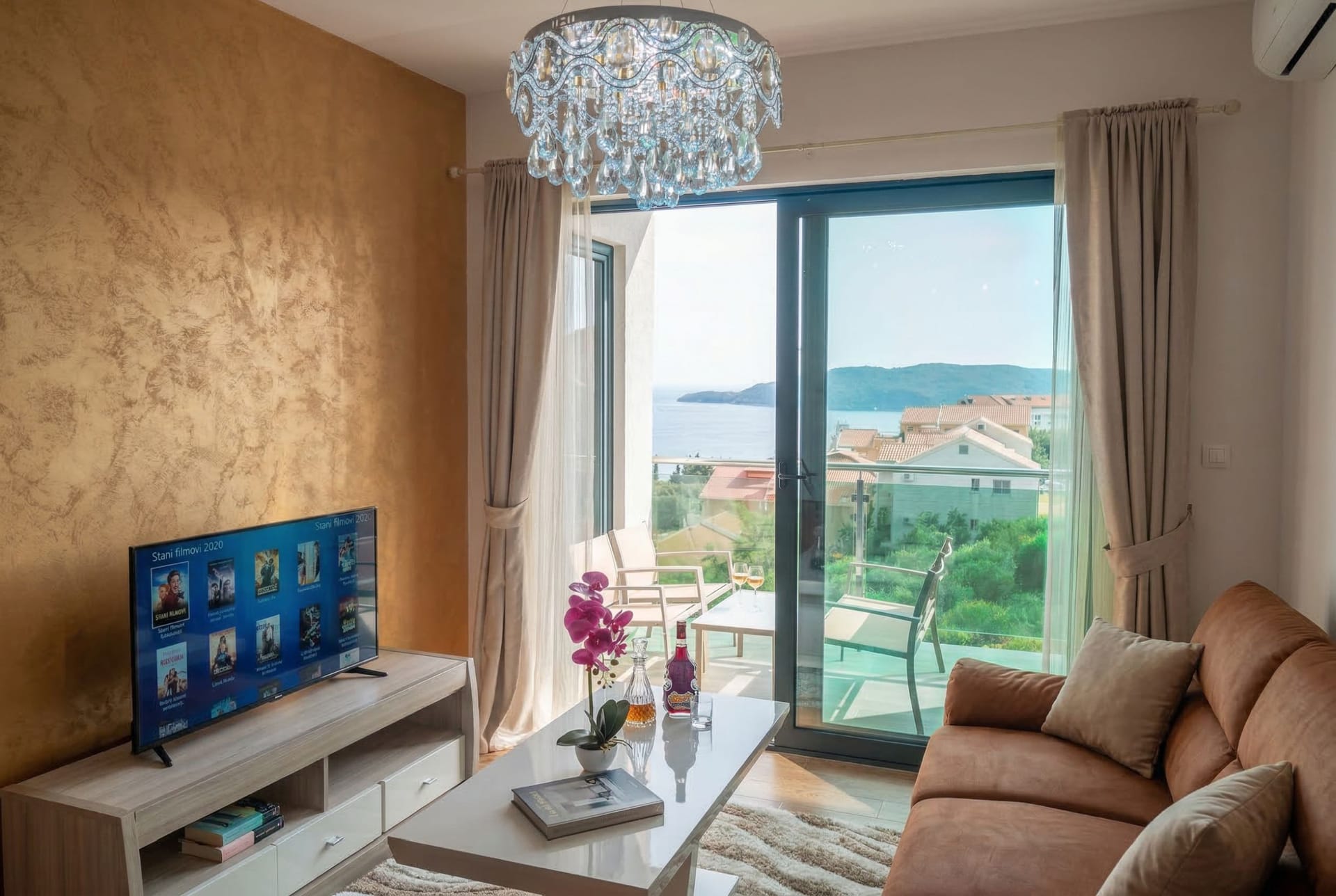 Dnevna soba u stanu u Bečićima, Crna Gora: pogled na more, balkon sa nameštajem, zlatni zid i kristalni luster.