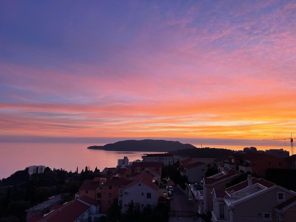 Panoramski pogled na more i zalazak sunca iz stana u Bečićima, Crna Gora: gradska izgradnja sa crvenim krovovima na obali.