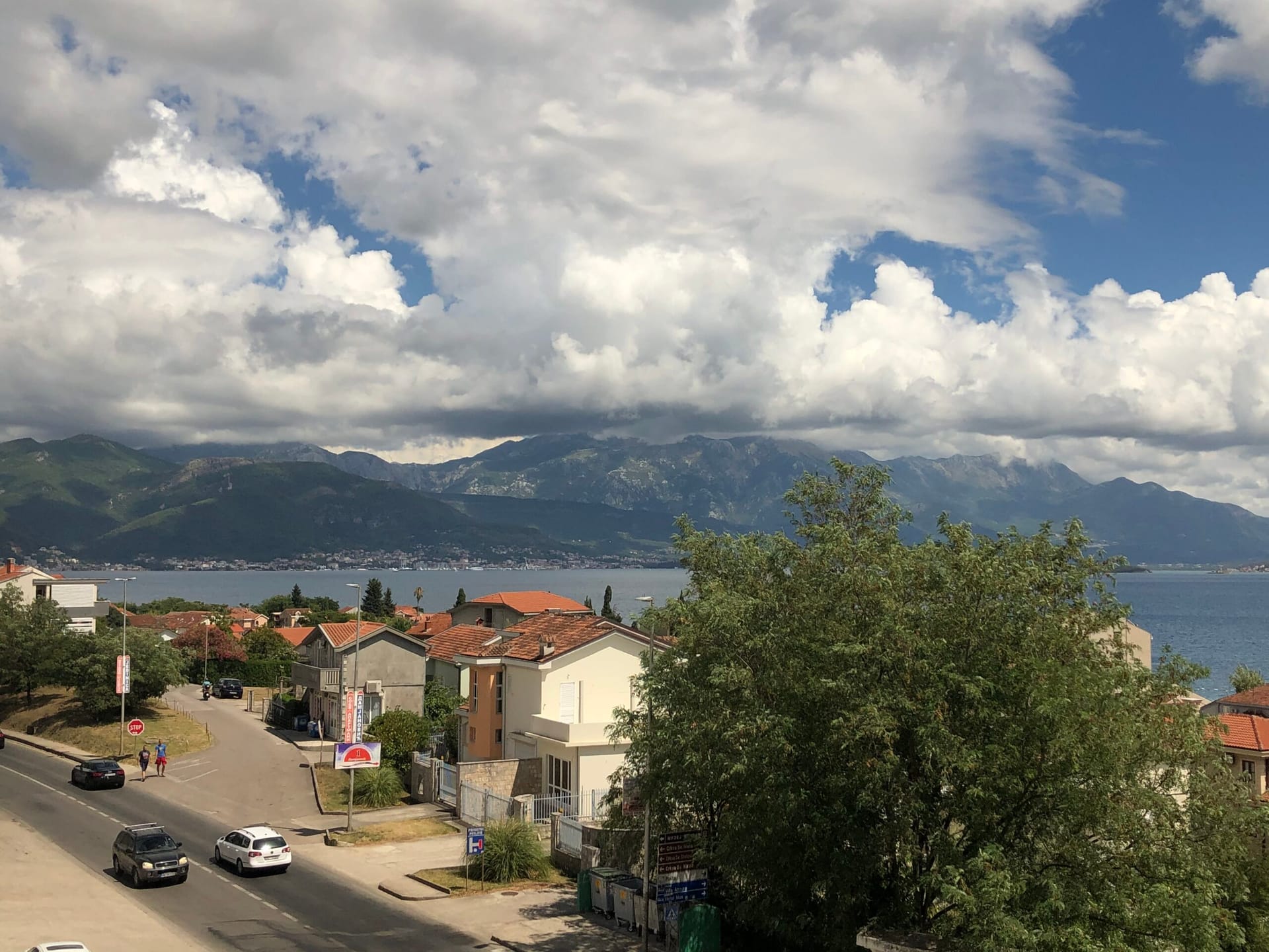 Panoramic sea and mountain view from an apartment in Baošići, Montenegro: coastal road, residential buildings, and cloudy sky.