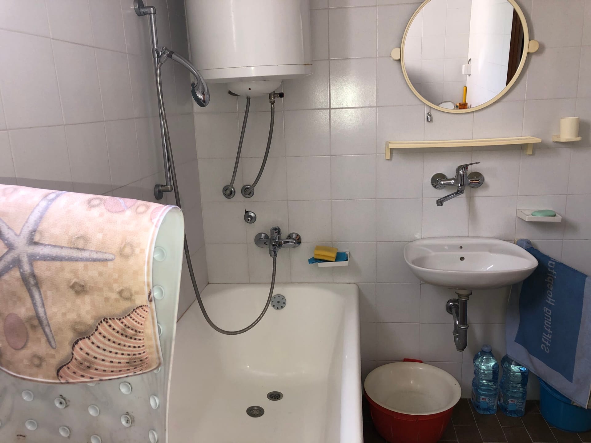 Bathroom in an apartment, Baošići, Montenegro: white tiles, bathtub, sink, water heater, and a round mirror on the wall.