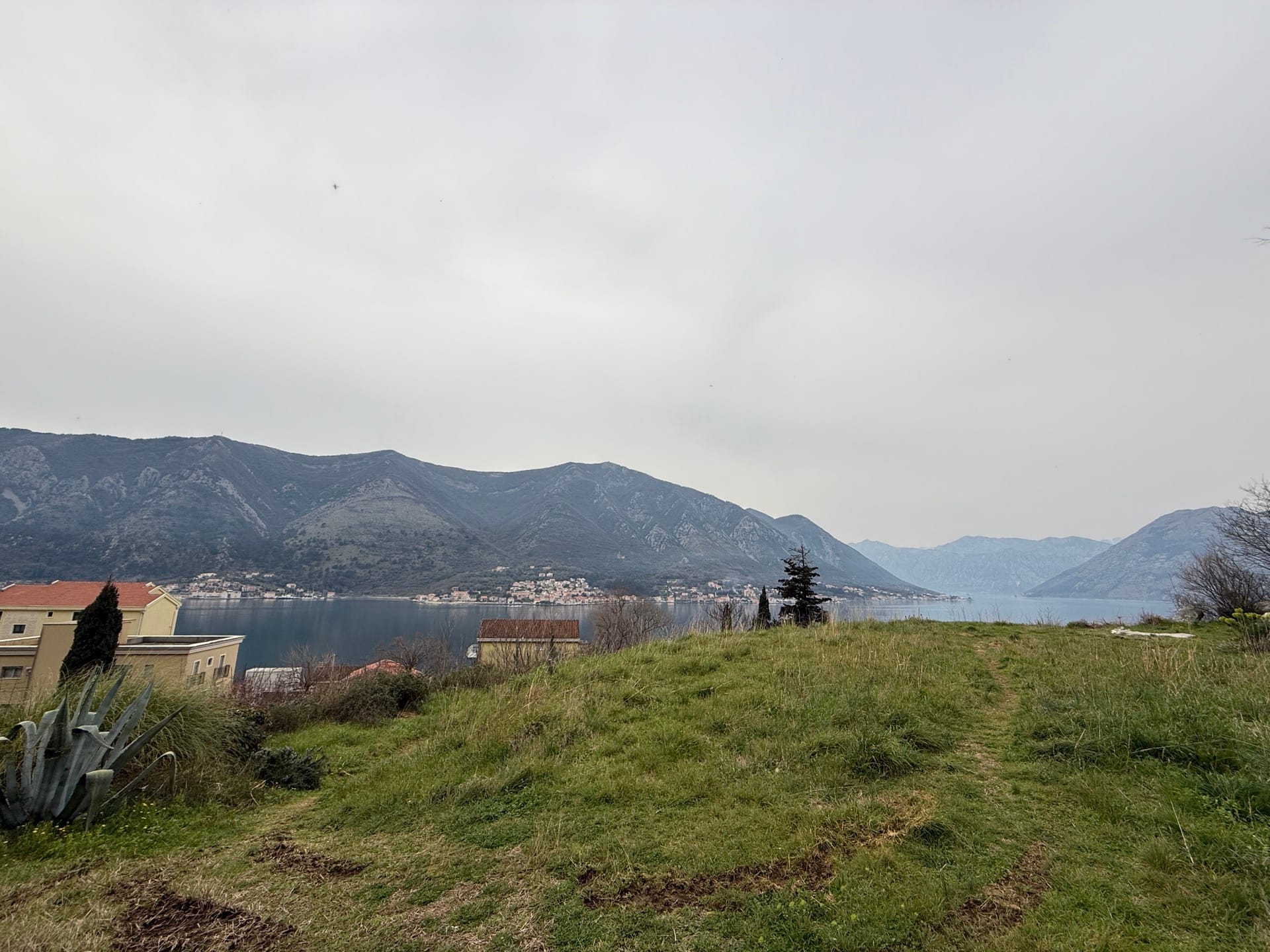Panoramic view of Kotor Bay and the mountains from Dobrota, Montenegro: a green plot with a house on the coast.